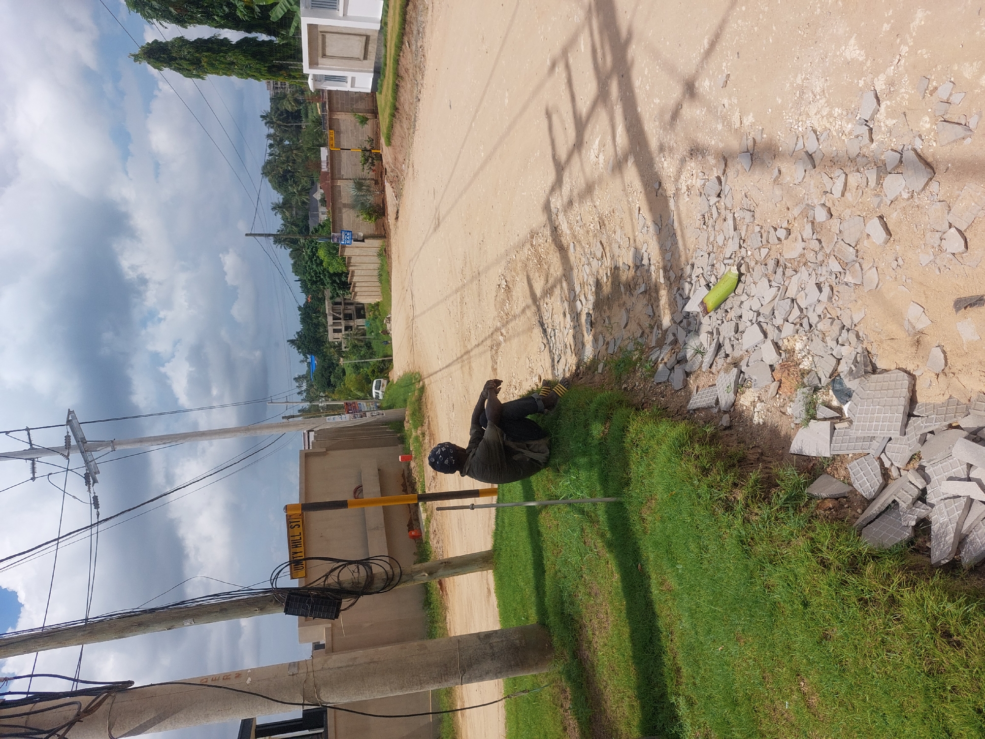 A man is sitting on the grass next to a dirt road in Dar es-Salaam, Tanzania. The road is lined with concrete debris and a green, cylindrical object. An electricity pole is on the other side of the grass. A sign that reads “Unity Hill Street” is attached to a building. The sky is cloudy, and there are trees and buildings in the background.