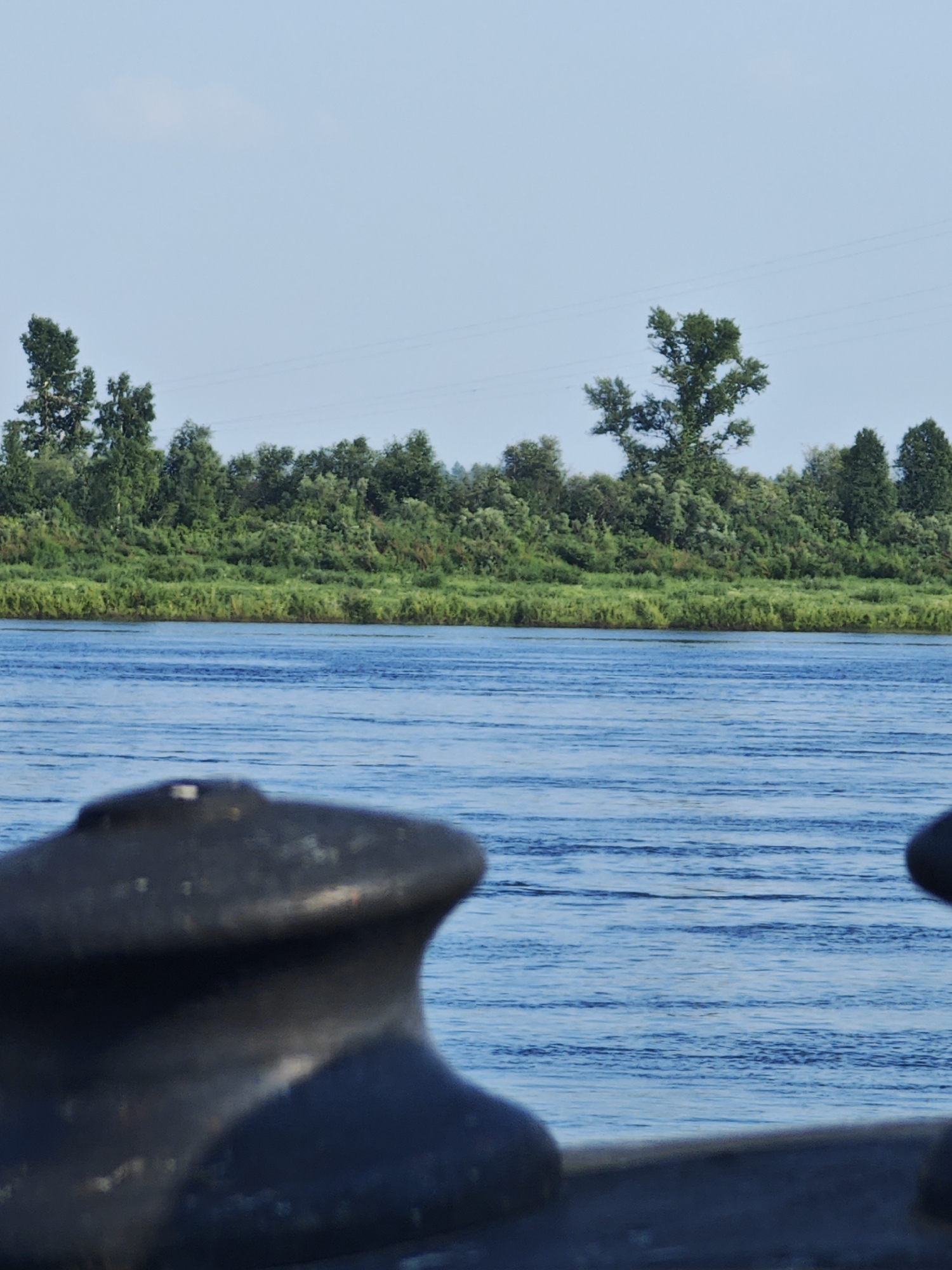 A close-up shot of a bollard on a boat, with a river and a tree-lined bank in the background. The bollard is dark and made of metal, with a rounded top. The river is blue and calm, with small ripples on the surface. The bank is covered in green trees and grass. The sky is clear and blue. The image appears to be taken on a sunny day in Верхнепашино, Russia.