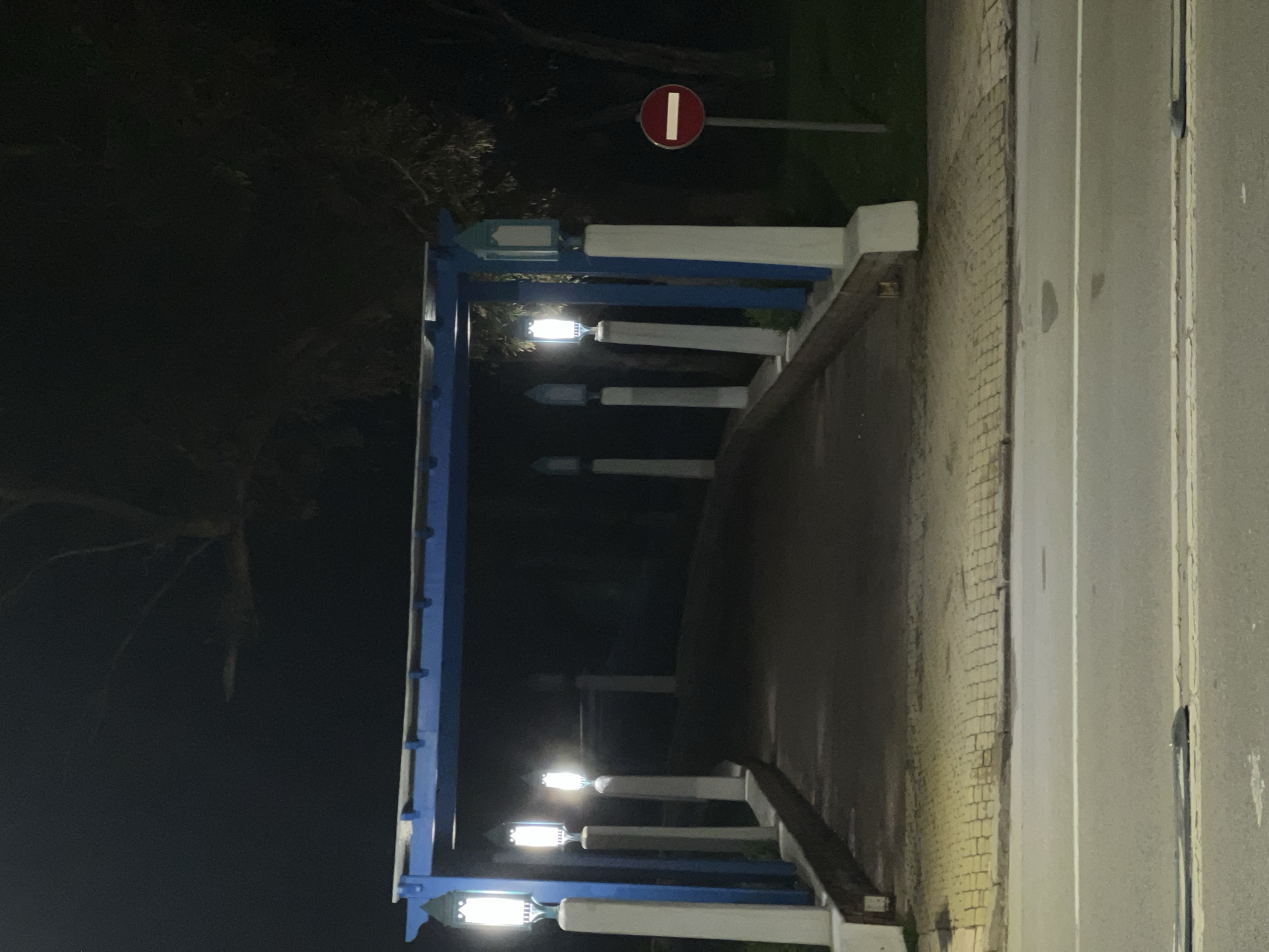 The image captures a pedestrian walkway or small bridge at night, illuminated by decorative lampposts. The structure features railings painted in white and a distinct light blue, adorned with five ornate lampposts that emit a bright, warm white light. This artificial lighting creates strong contrasts with the dark background, indicating nighttime.
To the right of the walkway, a paved road surface, likely asphalt, is visible, separated by a curb and a narrow strip of pavement. In the upper right portion of the scene, a red circular "No Entry" traffic sign with a white horizontal bar is prominently displayed on a pole. The background to the left is dark, with faint outlines of trees or dense vegetation, suggesting a natural or park-like setting adjacent to the path.
No people or activities are visible, presenting a quiet, static scene. The architectural style of the blue and white structure and the decorative lampposts suggest a public space, possibly a promenade or a themed area in M'Diq, Morocco. A textured wall, appearing to be made of brick, is visible in close-up views alongside the structure on the right. The overall impression is one of a well-maintained, aesthetically designed pathway under the night sky.