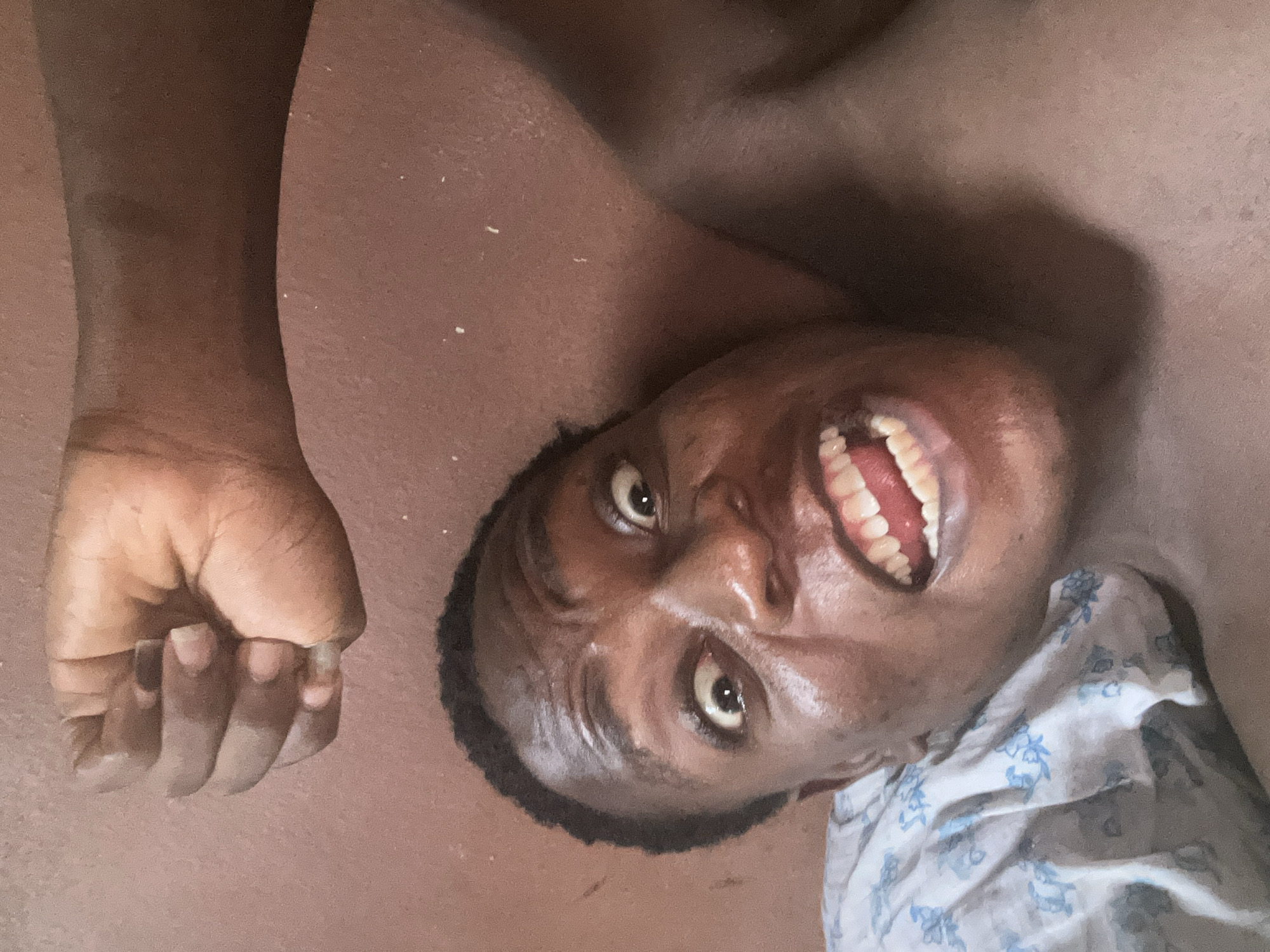 A person is laying on the floor, with their head tilted towards the camera. They have dark skin and are smiling widely, revealing their teeth. Their right hand is clenched into a fist and resting near their head. A floral patterned cloth is visible beneath the left side of their neck. The background is a plain, brown surface.