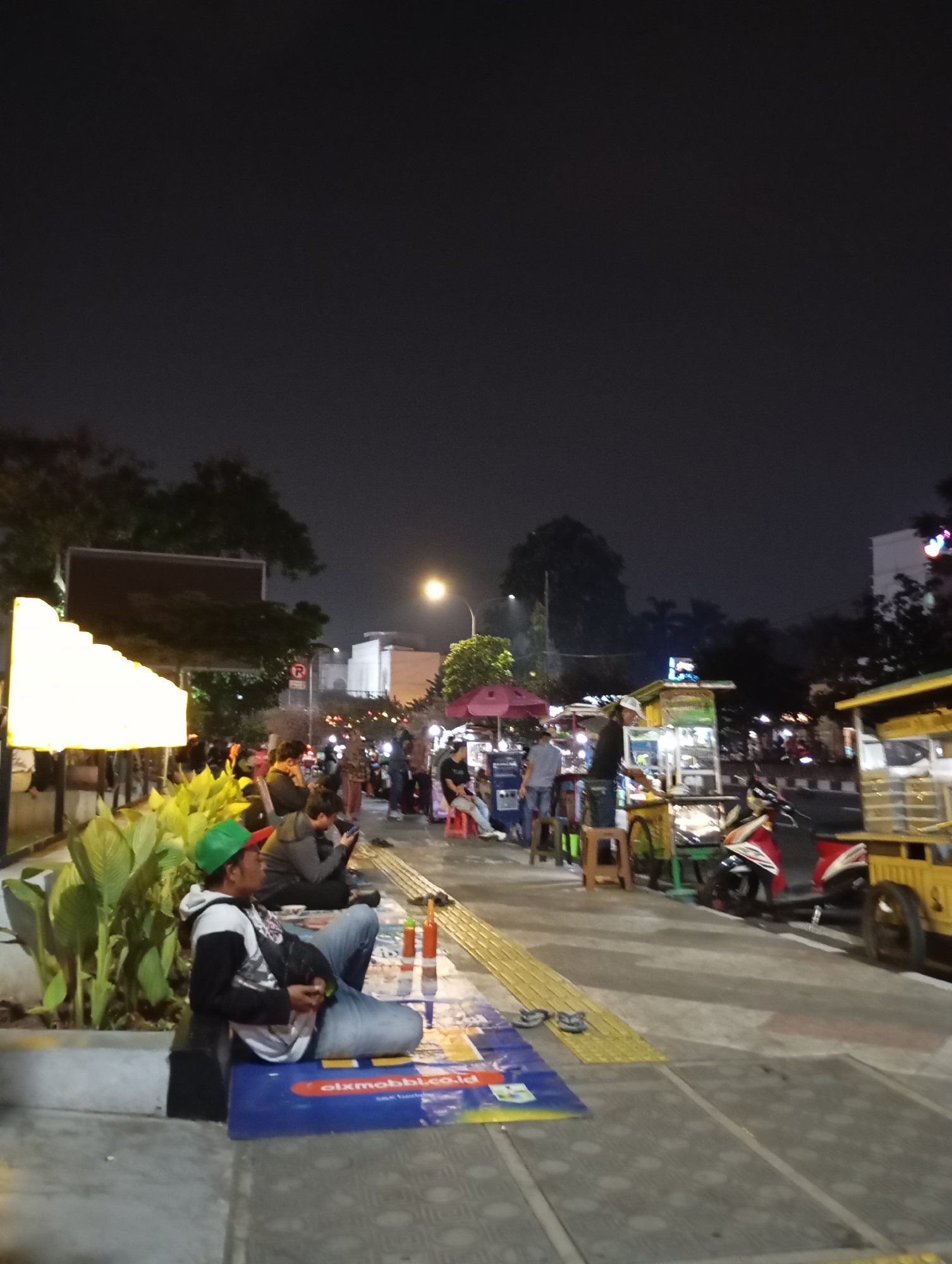 This is a nighttime scene in Depok, Indonesia, likely along a busy street or pedestrian walkway. A man is sitting on the sidewalk in the foreground, leaning against a planter. He is wearing a green cap, a hooded jacket, and jeans. In front of him, on the ground, is a blue mat with some writing on it and a pair of flip-flops. Several other people are visible in the background, some sitting at stalls or tables, and others walking along the street. There are food stalls and shops lining the walkway, illuminated by various lights. A motorcycle is parked near one of the stalls. The overall impression is that of a lively street scene at night, with people gathering and socializing at food vendors.