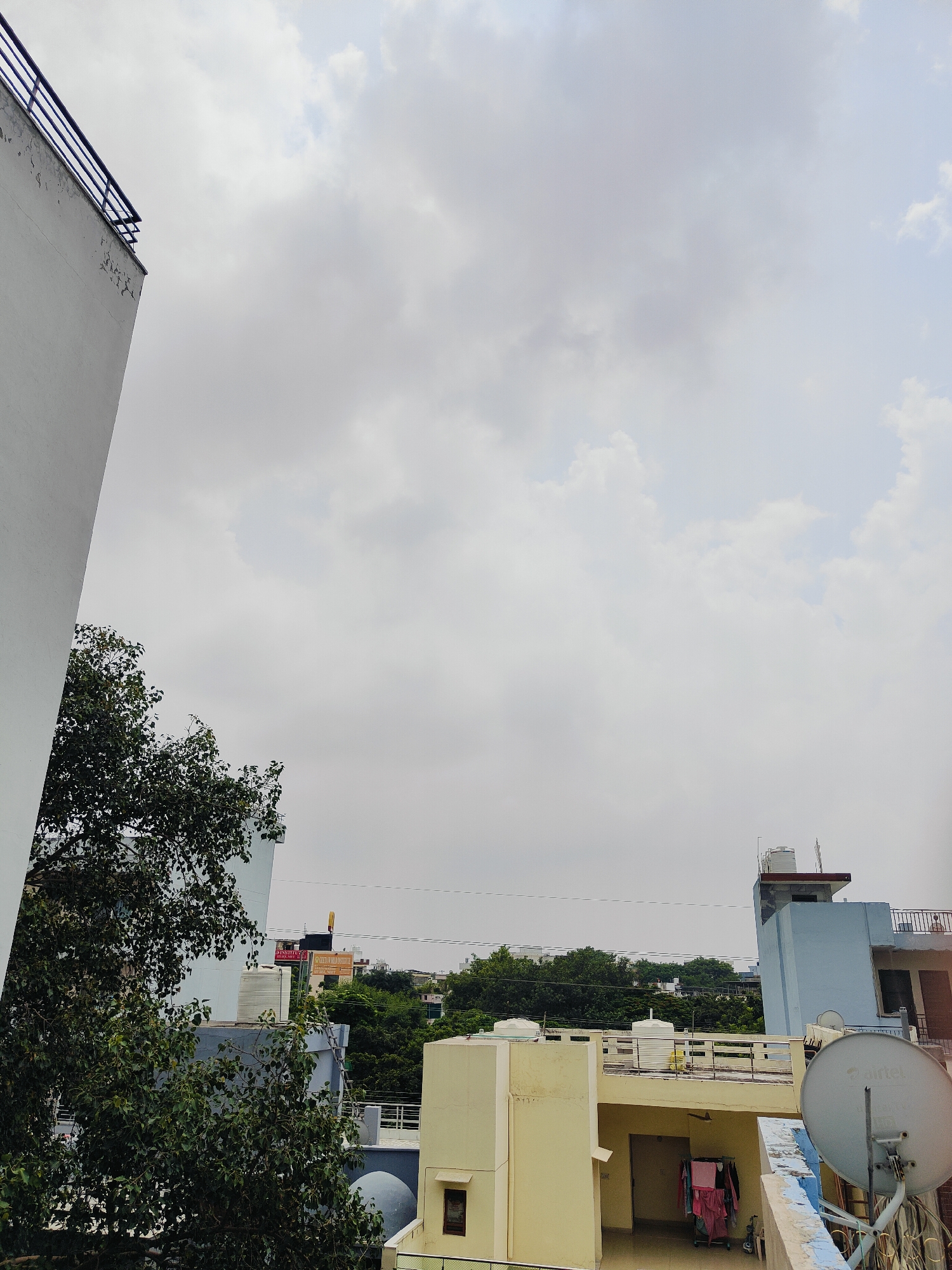 The image captures a view of rooftops in Gurgaon, India, under an overcast sky. A tall white building with a black railing dominates the left side of the frame, angled upwards. To the right, a lush green tree with dense foliage partially obscures a light blue building. Beyond that, several other buildings with flat roofs and varying shades of pastel colors are visible, interspersed with trees. One building features a water tank on its roof. A satellite dish, branded with "Airtel," is prominently positioned on a blue and white ledge in the lower right corner. Clothes are hanging on a rack on one of the lower rooftops, with a bicycle visible nearby. The overall scene depicts a dense urban environment with a mix of residential and possibly commercial buildings, characteristic of many parts of Gurgaon.