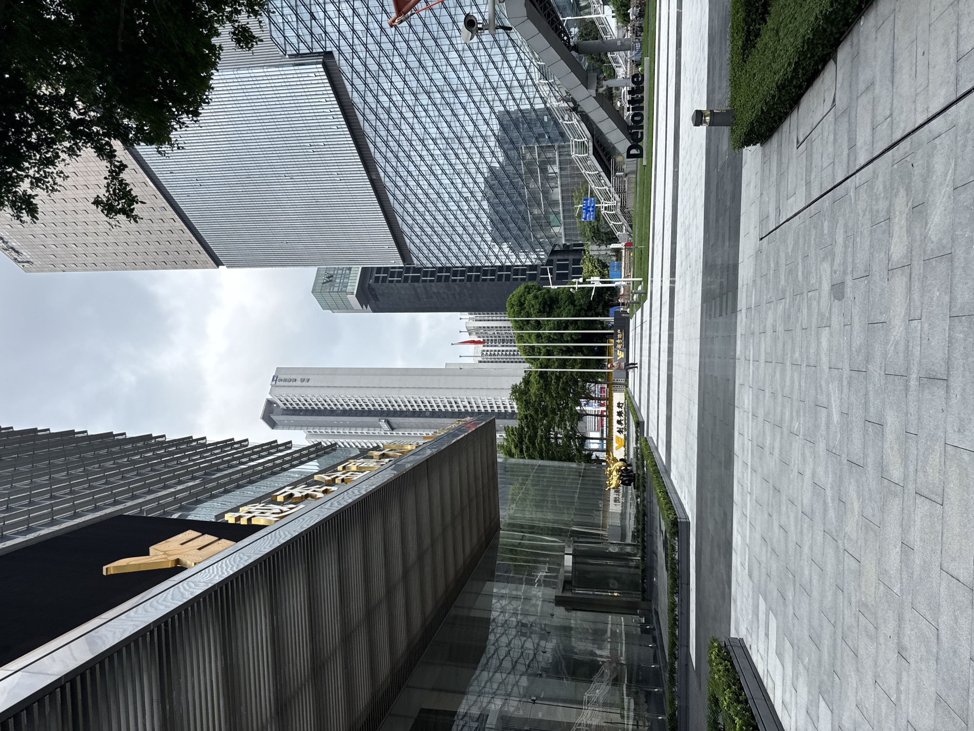 An exterior shot captures a modern urban environment in Tianhe District, China, likely on a cloudy day. The foreground features a wide, paved walkway with geometric patterns, bordered by manicured green hedges in raised planters. To the left, a large, cantilevered structure extends outwards, with its underside covered in horizontal metal slats. The facade of this building is primarily glass, reflecting the surroundings. Golden Chinese characters are visible on the building's facade.
In the mid-ground, a central plaza area is visible, with more paving stones and planters. A golden sculpture, possibly a dragon, is positioned within a planter. Further back, a street is lined with tall, modern skyscrapers. One prominent building has a glass facade with a distinctive diagonal pattern. A pedestrian bridge with a prominent "Deloitte" sign arches across the scene, leading to a raised walkway. A security camera is mounted on a pole near the bridge. Three red flags fly on poles in the distance, and several trees with green foliage are scattered throughout the area.
Two individuals are visible: one person is walking away from the camera on the left side of the walkway, and another person is walking towards the camera on the right side of the plaza. A yellow vehicle is parked near the building on the left. The sky is overcast, with muted light conditions.