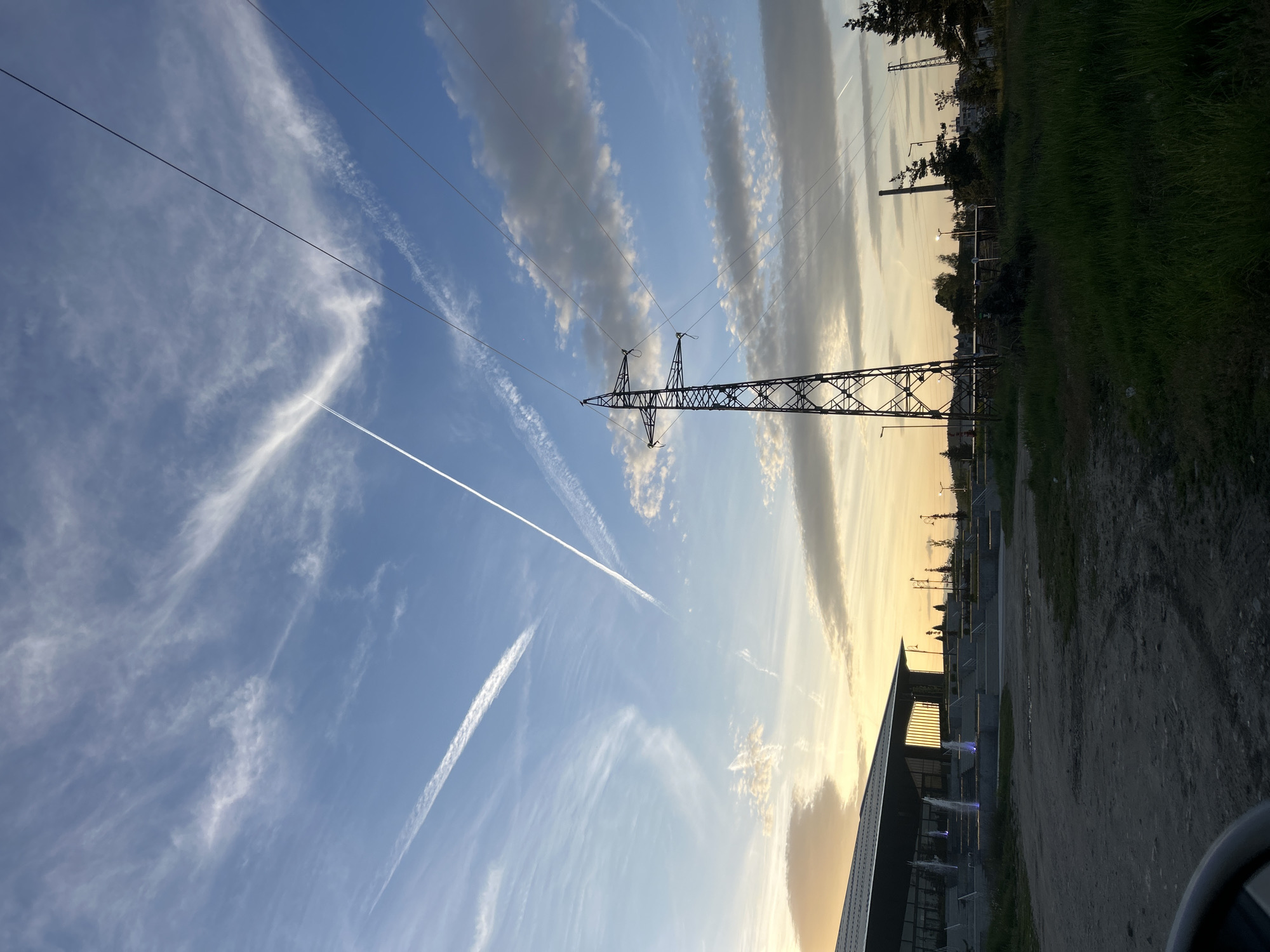 The image depicts a daytime scene in an unidentified location. An electrical transmission tower is centrally positioned, intersecting with power lines stretching across a partly cloudy sky. Two contrails bisect the sky diagonally, appearing in the lower-left quadrant. A building's roofline is visible at the bottom right, adjacent to a dirt or gravel road. Further into the background, trees are present. The sky appears blue and partially golden, suggesting it may be sunrise or sunset. No people are visible in the image.