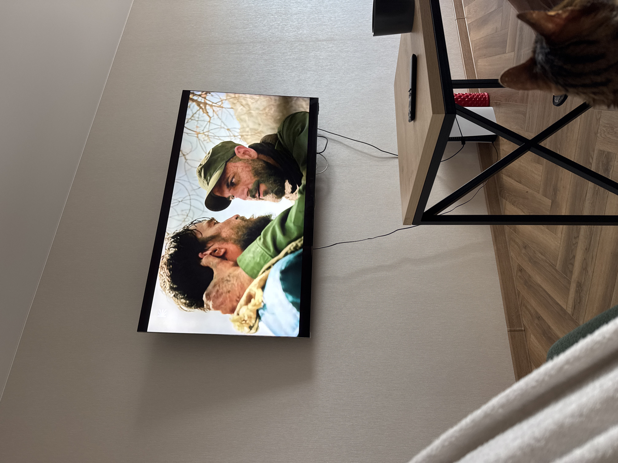 A television mounted on a gray wall displays a scene featuring two men, one wearing a green hat and the other with dark hair. To the right of the TV is a table with black metal legs and a light-colored wooden top. A cat is partially visible in the upper right corner. The floor appears to be wood-paneled, and the corner of a white blanket is visible in the lower right corner.