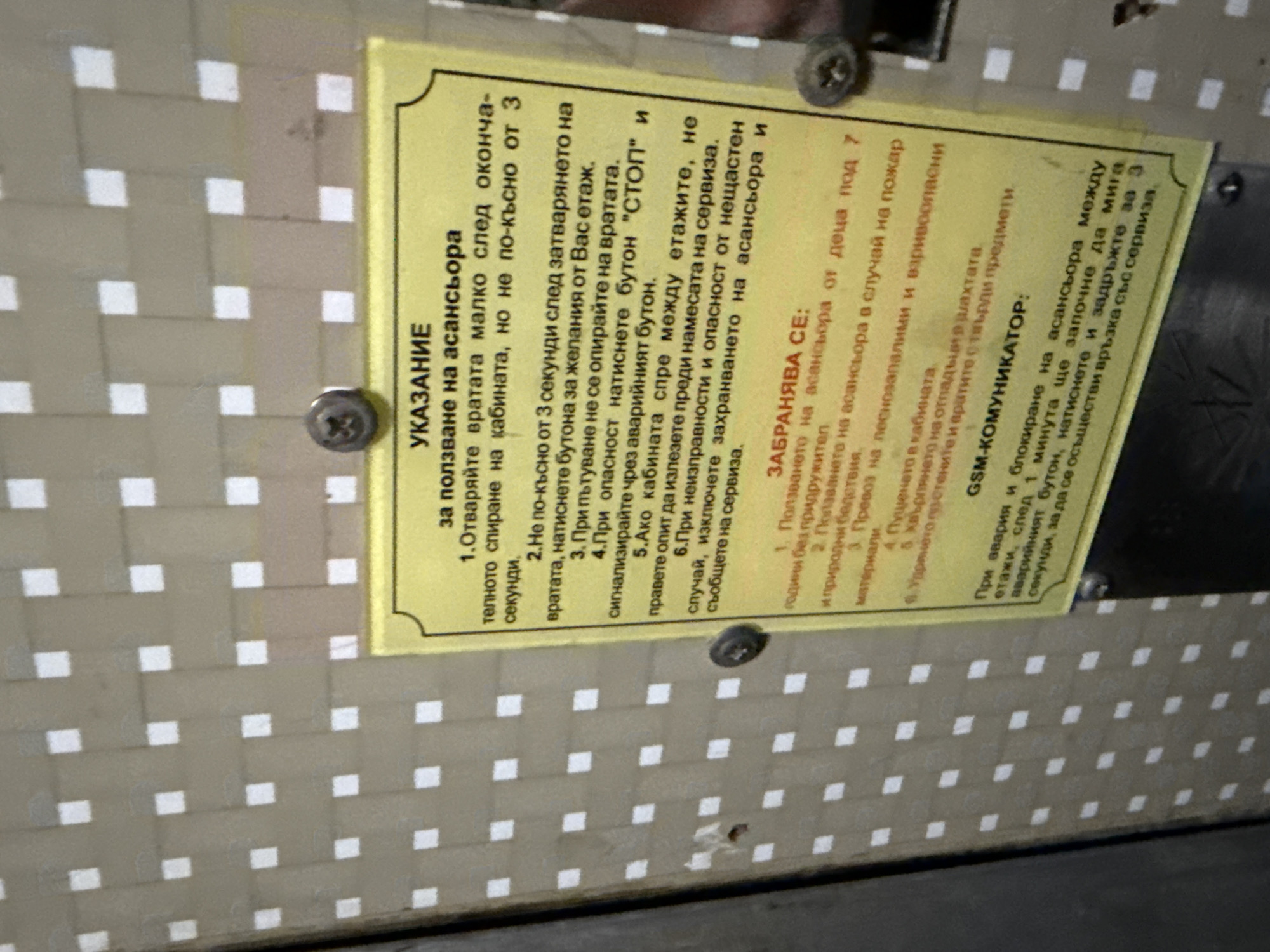 The image shows a set of instructions, written in Bulgarian, for using an elevator. The instructions cover normal operation, safety precautions, prohibited actions, and emergency communication via a GSM module. The sign is attached with screws to a wall that has a checkered pattern of light and dark squares.