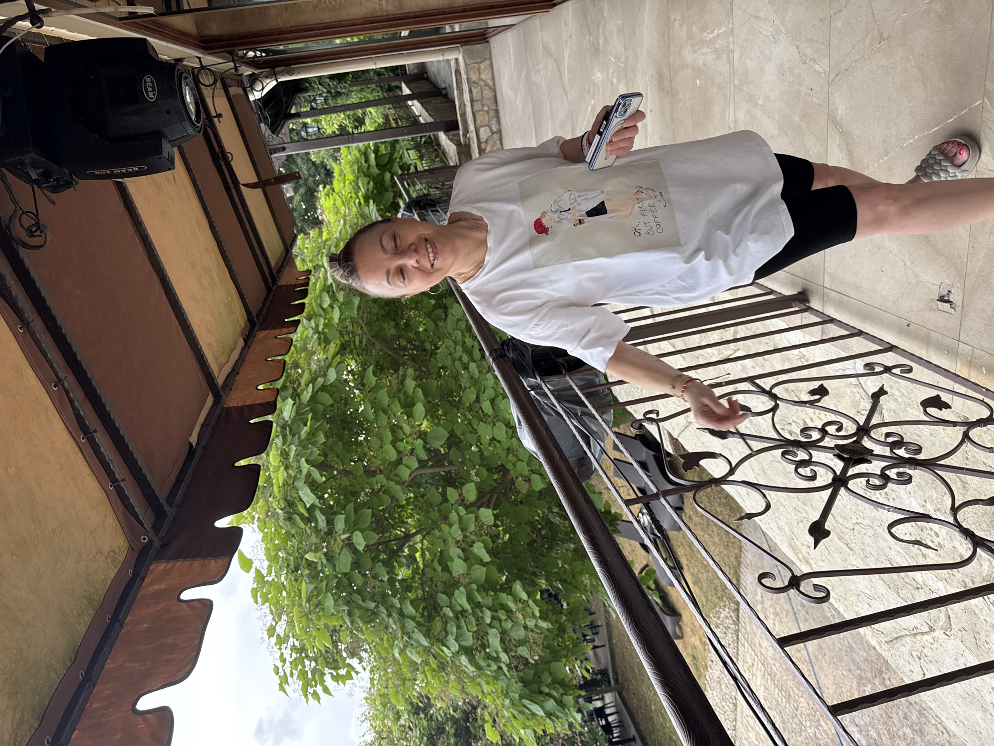 A woman stands on a stone patio, holding a smartphone. She is wearing a white t-shirt with a graphic print and black shorts. The t-shirt has an illustration of a person with a red hat and the text "OK. BUT FIRST COFFEE." She is smiling and looking towards the camera. In the background, there is lush green foliage and a metal railing with ornate scrollwork. Above, a section of a brown awning with a decorative edge is visible. To the left, a black spotlight fixture is mounted. The scene appears to be outdoors during the daytime, with a bright, overcast sky. The overall impression is casual and relaxed. The location is Sredets, Bulgaria.