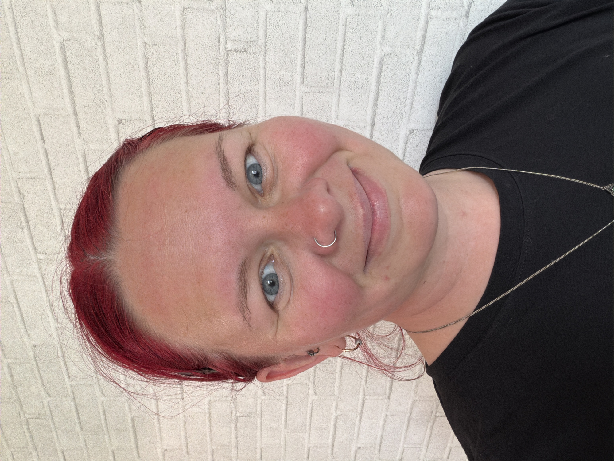 A woman with red hair, blue eyes, and a nose ring is facing the camera and smiling. She is wearing a black shirt and a silver necklace. The background is a white brick wall. It is not possible to confirm that this image was taken in the city of Chaikovsky, Russia from the visual information available.