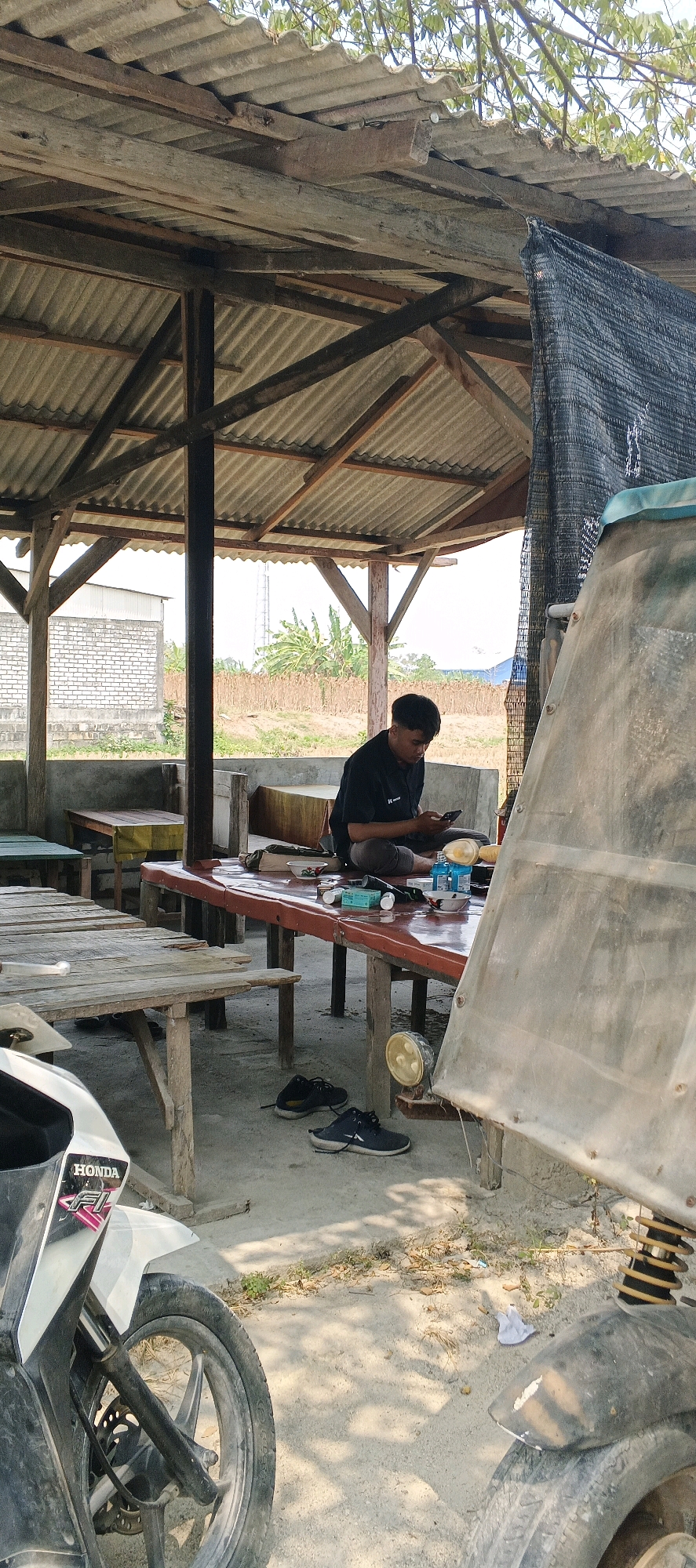 The image depicts an outdoor seating area, likely a roadside eatery or rest stop in Sonoadi, Indonesia. A young man wearing a black t-shirt is seated at a long, red-topped table, engrossed in his mobile phone. Scattered on the table are various items including a small bowl of food, a plastic container, a small bottle, and what appear to be packets of cigarettes. Two pairs of dark-colored shoes are placed on the dusty ground near the table. In the foreground, the front portion of a white Honda motorcycle is visible, suggesting recent arrival or departure. The seating area is covered by a rustic, corrugated metal roof supported by wooden beams, with additional netting on one side. Behind the table, a low brick wall and further out, a field with scattered vegetation and distant buildings are visible, indicating a semi-rural or peri-urban setting. The lighting suggests it is daytime, with sunlight filtering through the overhead structure.