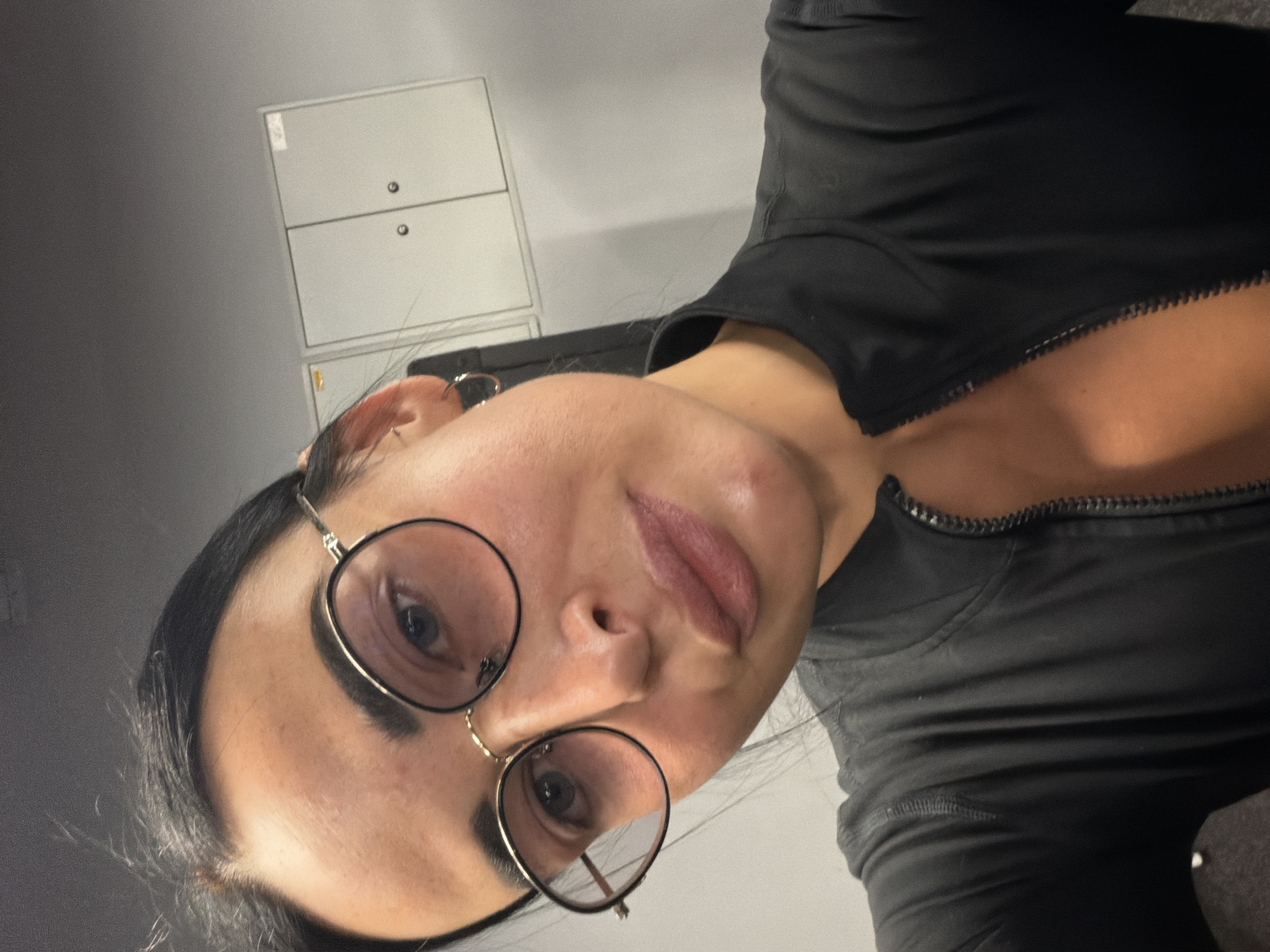 The image is a close-up selfie featuring a person, likely a woman, with dark hair and eyebrows, wearing round glasses with gold-toned frames. Her lips are slightly parted, showing a subtle pink lipstick. A hoop earring is visible in her left ear. She is wearing a black top, possibly activewear, with a prominent metallic zipper running down the front or side. The photograph is taken from a high angle, looking downwards, and is rotated 90 degrees counter-clockwise.
The background is simple, consisting of a plain grey wall. Mounted on this wall are two rectangular, light-grey cabinets or lockers, stacked vertically. The upper cabinet has a small white label or sticker, and the lower one has a small gold-colored sticker. Both cabinets feature small black circular details, likely locks or handles. The setting appears to be an indoor, utilitarian space, such as a locker room or a changing area. While the location context is Saint Petersburg, Russia, no specific visual elements in the image confirm this geographical detail.
The lighting is moderate, creating some soft shadows on her face and enhancing the texture of her top. Her expression is neutral or slightly pensive. No discernible text is visible on the person, and the text on the cabinet labels is too small to read. The overall impression is an intimate, slightly tilted portrait within a functional environment.