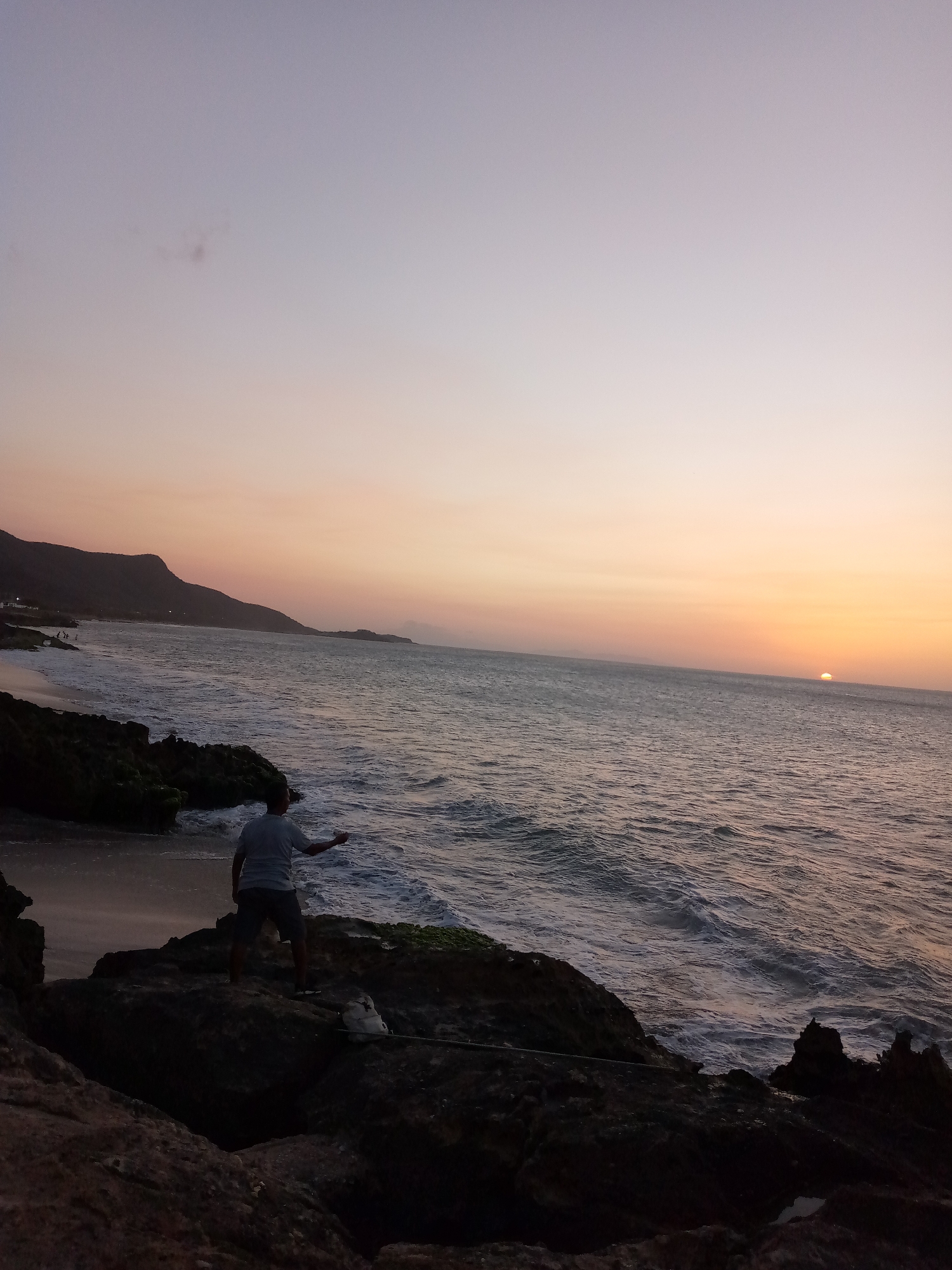 The image captures a sunset scene at Juan Griego, Venezuela. An adult male stands on a rocky outcrop, facing the ocean, possibly fishing. The sun is setting on the horizon, casting a warm orange glow on the water and sky. The coastline is visible in the background with mountains. Waves crash against the rocks. There's some fishing equipment at the base of the rocks where the man is standing. The sky transitions from a pale purple at the top to a warm orange near the horizon. The lighting suggests it is either sunrise or sunset.