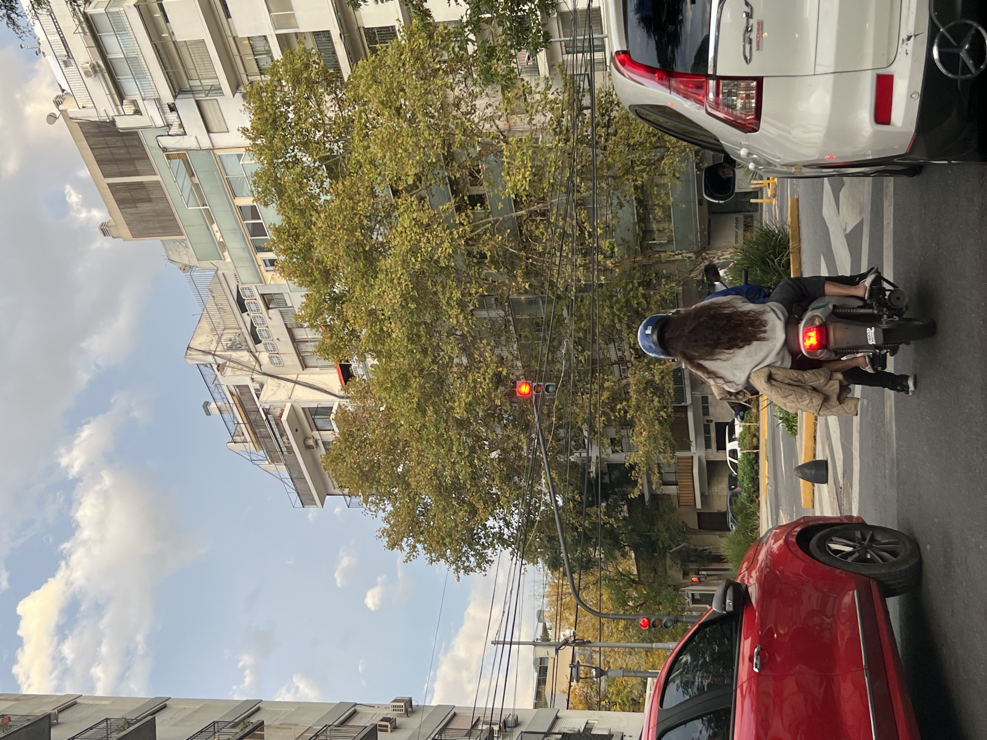 A street view in Buenos Aires, Argentina, showing a person with long hair riding a moped in traffic. The person is wearing a blue helmet. A white car is directly in front of the moped, and a red car is behind. A tree with green leaves is in the background, partially obscuring buildings. The traffic lights are red. The sky is blue with scattered clouds. Power lines are visible overhead. Buildings with balconies are on either side of the street.