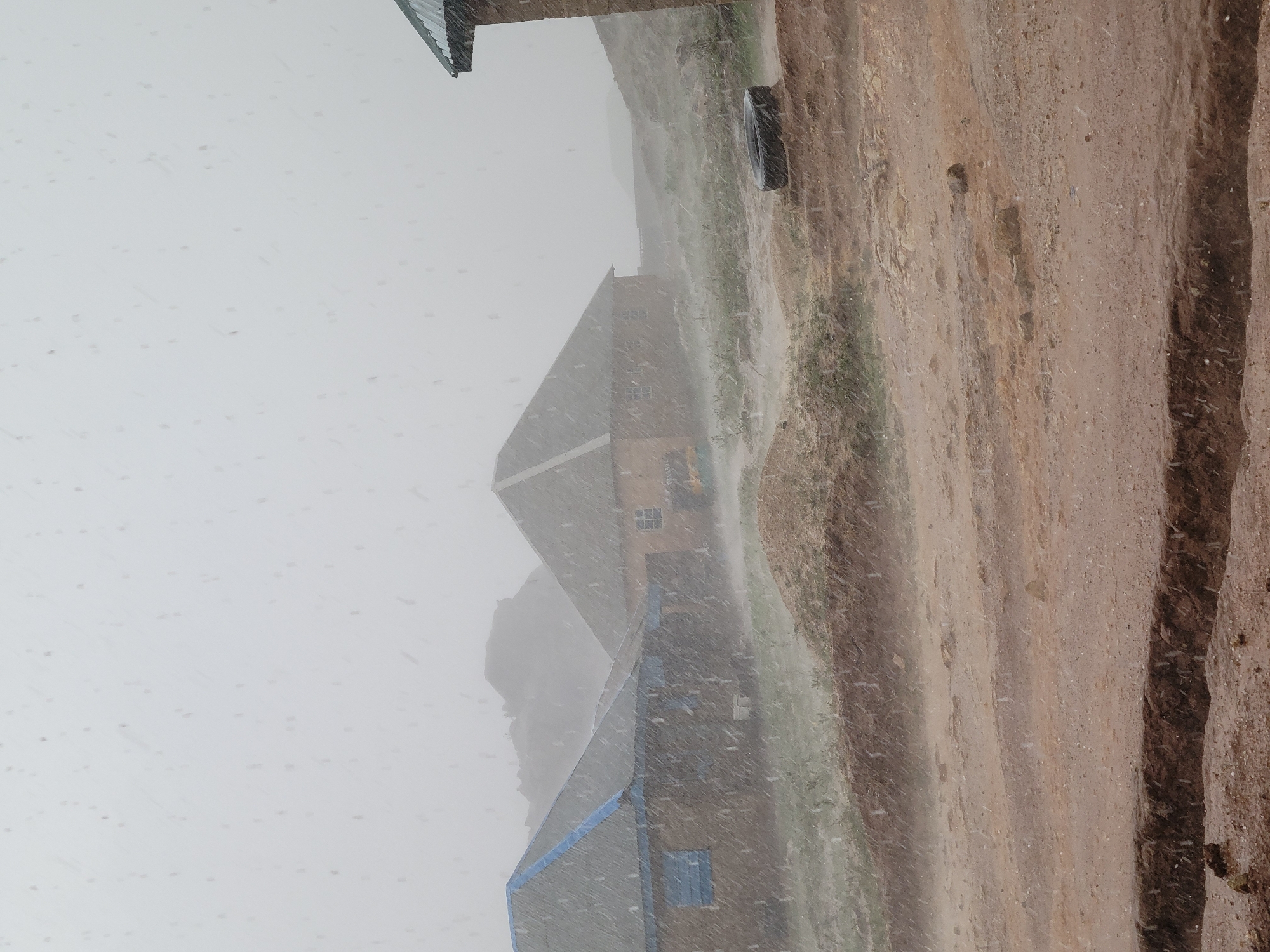 The image shows a heavy rainfall event in Barakin Akawo, Nigeria. Several buildings are visible, obscured by the rain. A dirt or gravel road runs alongside the buildings, and the ground is saturated with water. The overall visibility is low. No people are visible in the image.
