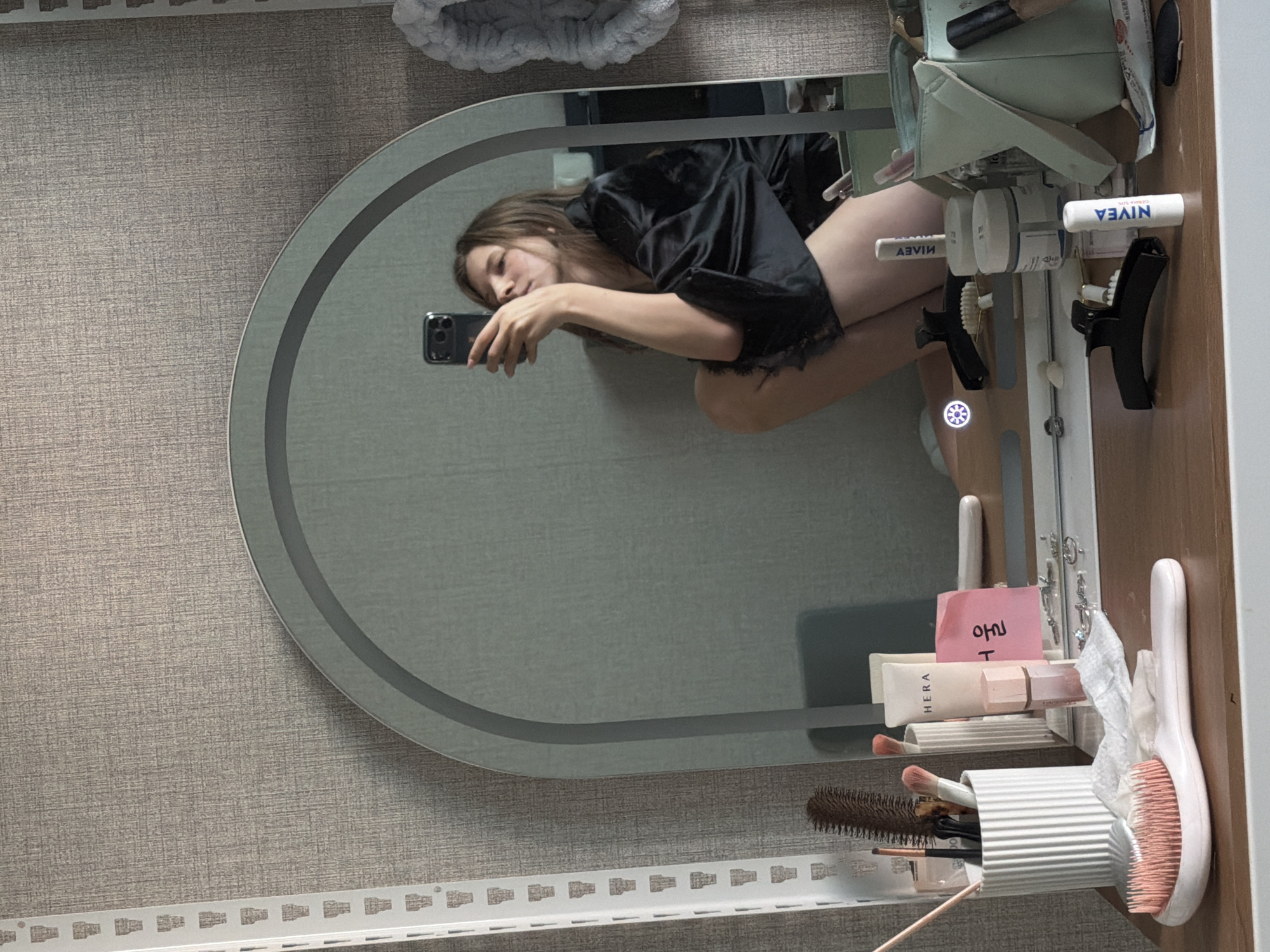 A young woman is taking a mirror selfie in what appears to be a bedroom or vanity area. She is wearing a black satin robe with lace trim and is crouching down, holding up her phone. The mirror is large and has an arched top with integrated lighting. On the vanity table below the mirror are various beauty products, including makeup brushes in a white holder, a tube of HERA sunscreen, a small pink bottle, a Nivea lip balm, and other containers. A light blue makeup bag is also visible. In the background, the wall has a textured wallpaper. To the left of the mirror is a white metal shelving unit. The scene is illuminated by the mirror's lights and possibly ambient room lighting. The overall impression is of a personal grooming space. The location is identified as Dongducheon-si, South Korea.
