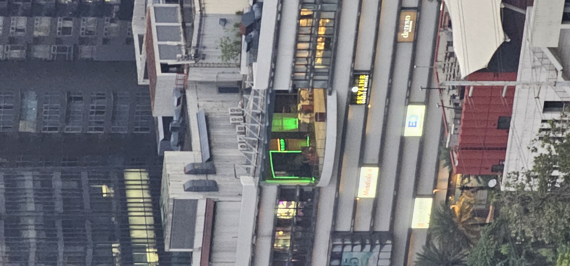 The image shows a multi-story building in Makati City, Philippines. The building appears to be a commercial or mixed-use development with several businesses advertised on its facade. Visible signs include "Mendiola's," "SKYLINE SPORTS & SOCIAL," "ALTOS TOPTI," and "denovo." There is a section of the building with green neon lighting and glass windows, suggesting a restaurant or lounge area. Other architectural features include various windows, air conditioning units, and what appears to be construction or renovation work on some of the rooftops, indicated by red tarpaulins. The background features taller office buildings typical of Makati's skyline. The overall scene depicts an urban environment with commercial activity.