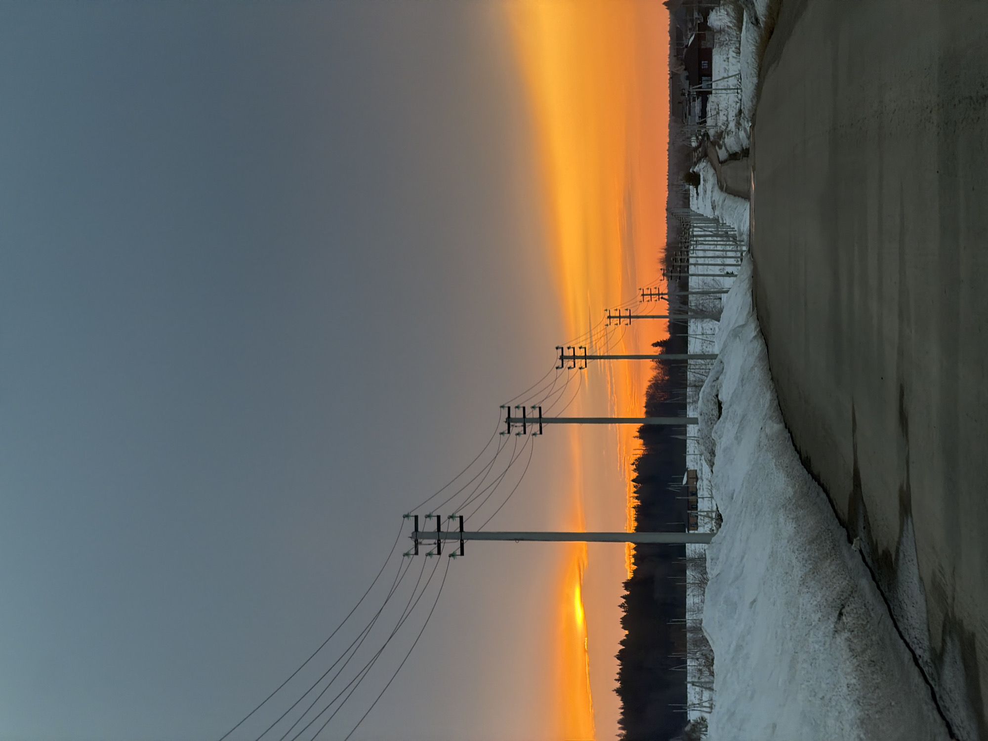 The image shows a sunset over a road in a rural area. The sky is divided, with a dark gray-blue on the left and a vibrant orange on the right, with the orange hue appearing to be reflecting off low-lying clouds. Power lines and poles run along the left side of the road, silhouetted against the sky. On the right side of the road, there is a snow-covered embankment, and in the distance, there are buildings or houses partially obscured by trees. It is not possible to confirm the exact location in the city of Izhevsk, Russia, without additional information.