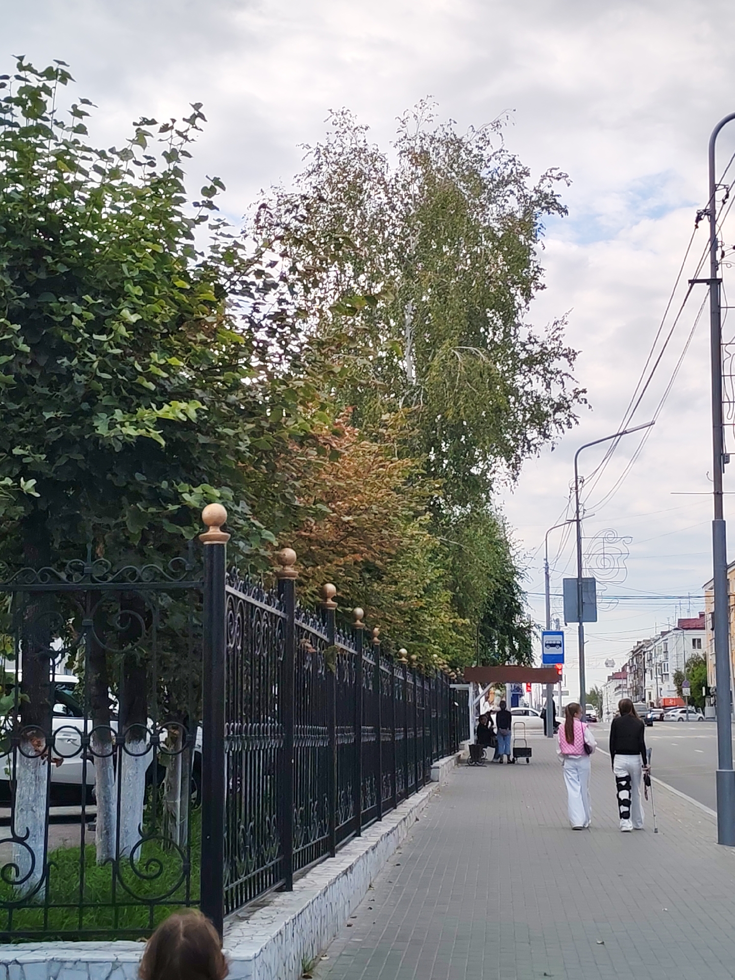 The image depicts an outdoor scene on a city sidewalk. In the foreground, a black wrought-iron fence with decorative gold finials runs along the left side of the frame, backed by green grass and trees. A white tiled wall borders the sidewalk. Two young women are walking away from the camera on the sidewalk. The woman on the right is wearing a black jacket and white pants, and has a brace on her left leg and is using a crutch. The woman on the left is wearing a pink vest over a white outfit. In the background, there is a bus stop with a shelter and a bus stop sign. Further down the sidewalk, several people are visible near the bus stop, including a person sitting on a bench and another pushing a cart. A white car is parked behind the fence on the left. The street runs parallel to the sidewalk, with cars visible in the distance. Buildings line the opposite side of the street. The sky is overcast with some clouds.