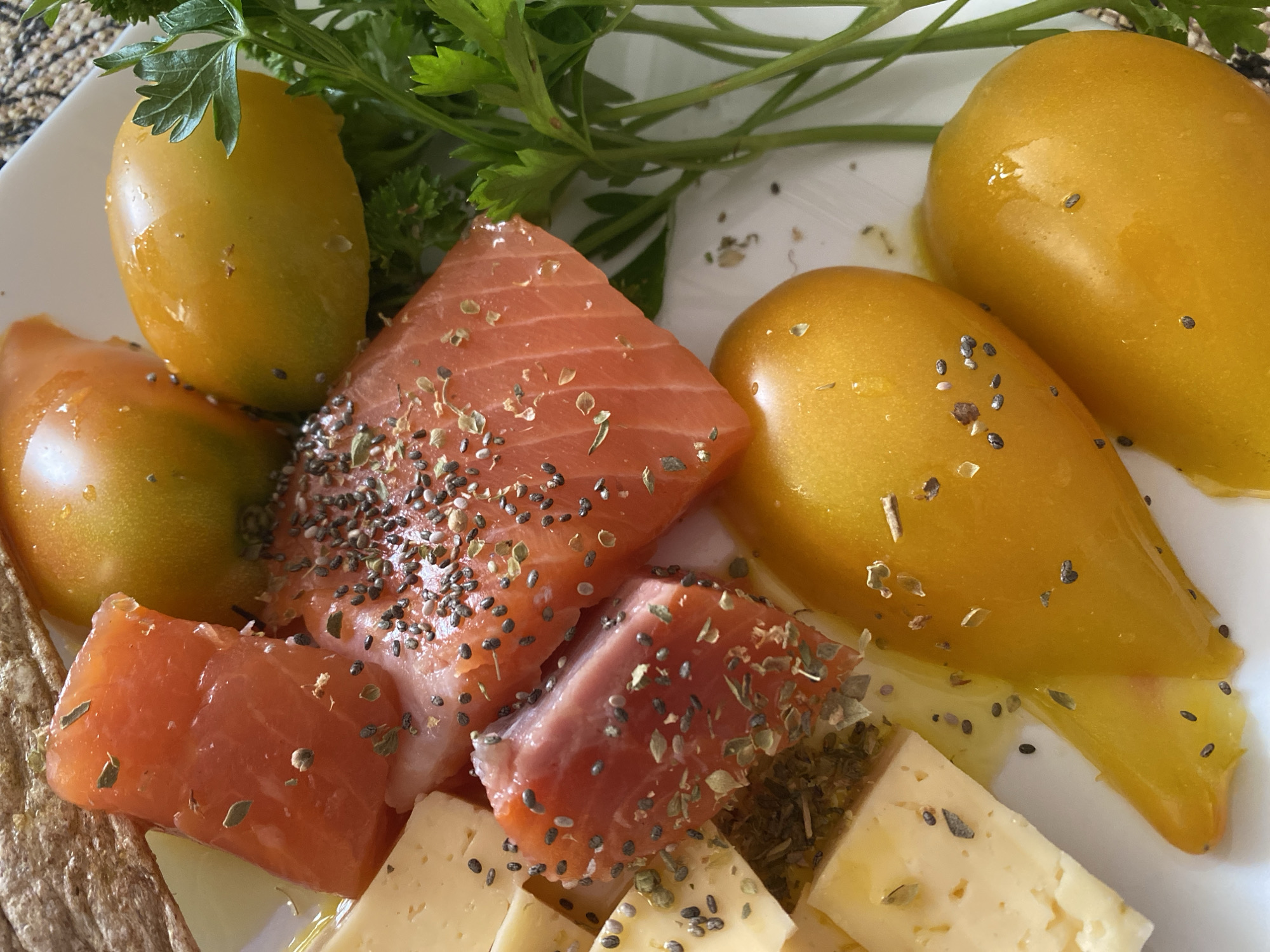 A close-up, overhead shot of a white plate filled with food. The plate contains pieces of raw salmon, cubed cheese, yellow tomatoes, and a garnish of parsley. The salmon is seasoned with what appears to be chia seeds and dried herbs. The cheese is pale yellow and appears to be a firm variety. The tomatoes are halved and glistening, also with some visible seasoning. The parsley is fresh and green. The overall presentation suggests a healthy meal or appetizer. The location in Lida, Belarus is not discernible from the image itself.