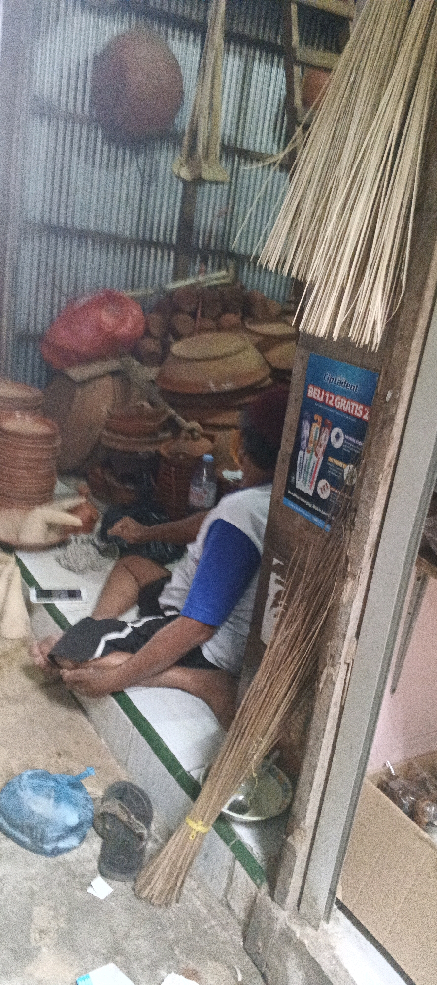 In a dimly lit interior, possibly a workshop or storage area, a person sits on a white and green elevated surface, resting their feet on what appears to be a dark fabric. The person is wearing a white shirt with blue sleeves and black shorts with white stripes. A mobile phone is placed on the surface next to them. The surrounding environment is cluttered with various objects.
To the left, a stack of terracotta-colored plates is visible. Behind them, large terracotta pots and a red plastic bag are piled up. A bundle of dried grass or reeds hangs from the ceiling.
To the right of the person, a wooden plank leans against the wall, adorned with a poster for "Cipladent" toothpaste, advertising a "Beli 12 Gratis 2" (Buy 12 Get 2 Free) promotion. A broom made of dried reeds is tied with a yellow band and rests against the plank. In the background, more terracotta pots of various sizes are stacked, along with a water bottle.
The walls appear to be corrugated metal, suggesting a rustic or industrial setting. The floor is concrete. The overall impression is one of a working space filled with handcrafted items and materials. The presence of the poster suggests this location may be near a commercial area or that the owner is actively engaged in sales or marketing.
This scene is located in Sekaran, Indonesia.