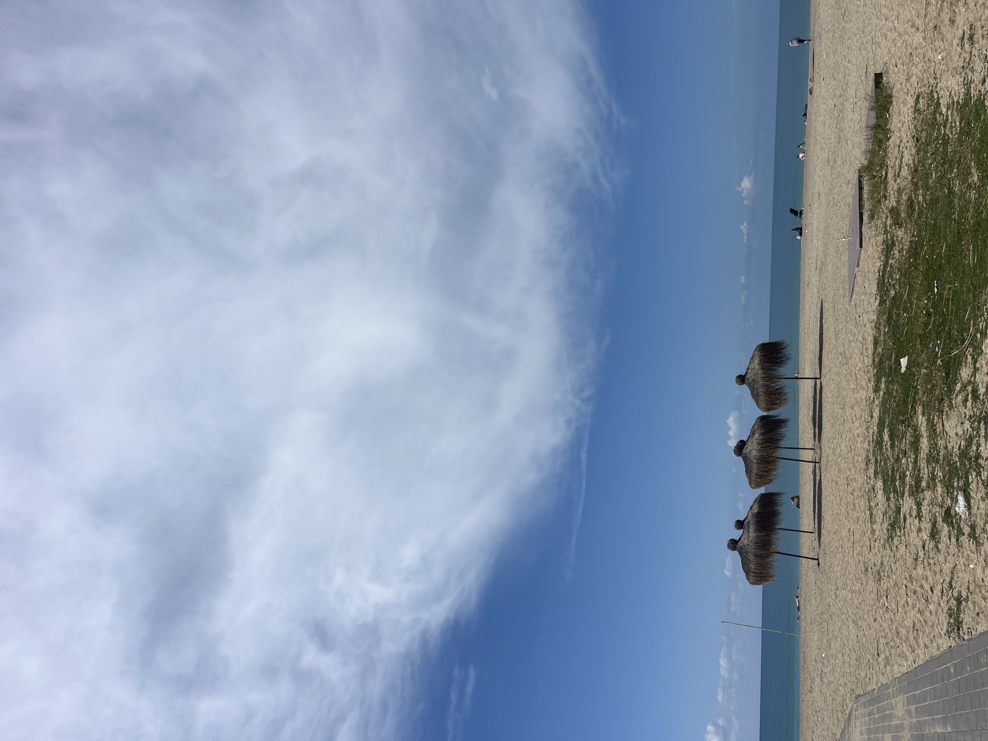 The image shows a beach scene in Alanya, Turkey. The view is oriented to show the coastline running from bottom to top of the image. Three thatched umbrellas are aligned along the beach near the water's edge. Patches of green vegetation grow in the sand on the right side of the image. People are visible on the beach. The sky is partly cloudy. The sea is calm and blue.