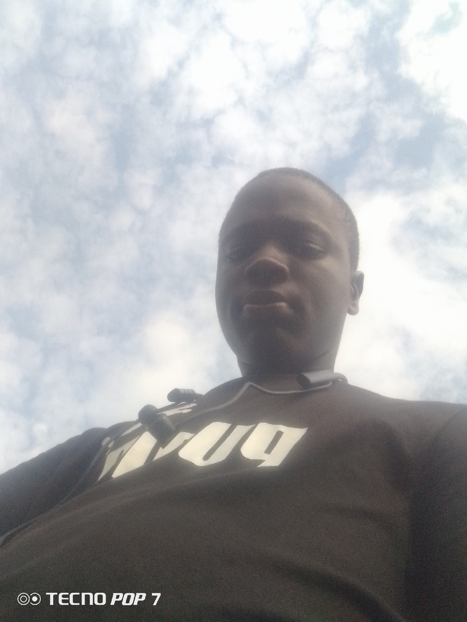 A low-angle, close-up selfie shows a young Black male from the chest up against a cloudy sky. The subject's head is tilted back, and his gaze is directed upwards. He is wearing a black t-shirt with large, stylized white lettering across the chest, partially obscured by the camera angle. A dark grey pair of earbuds with black tips rests around his neck. The lower portion of the image shows the text "TECNO POP 7" in white font. The image appears to have been taken outdoors in Eleweran, Nigeria, given the watermark.