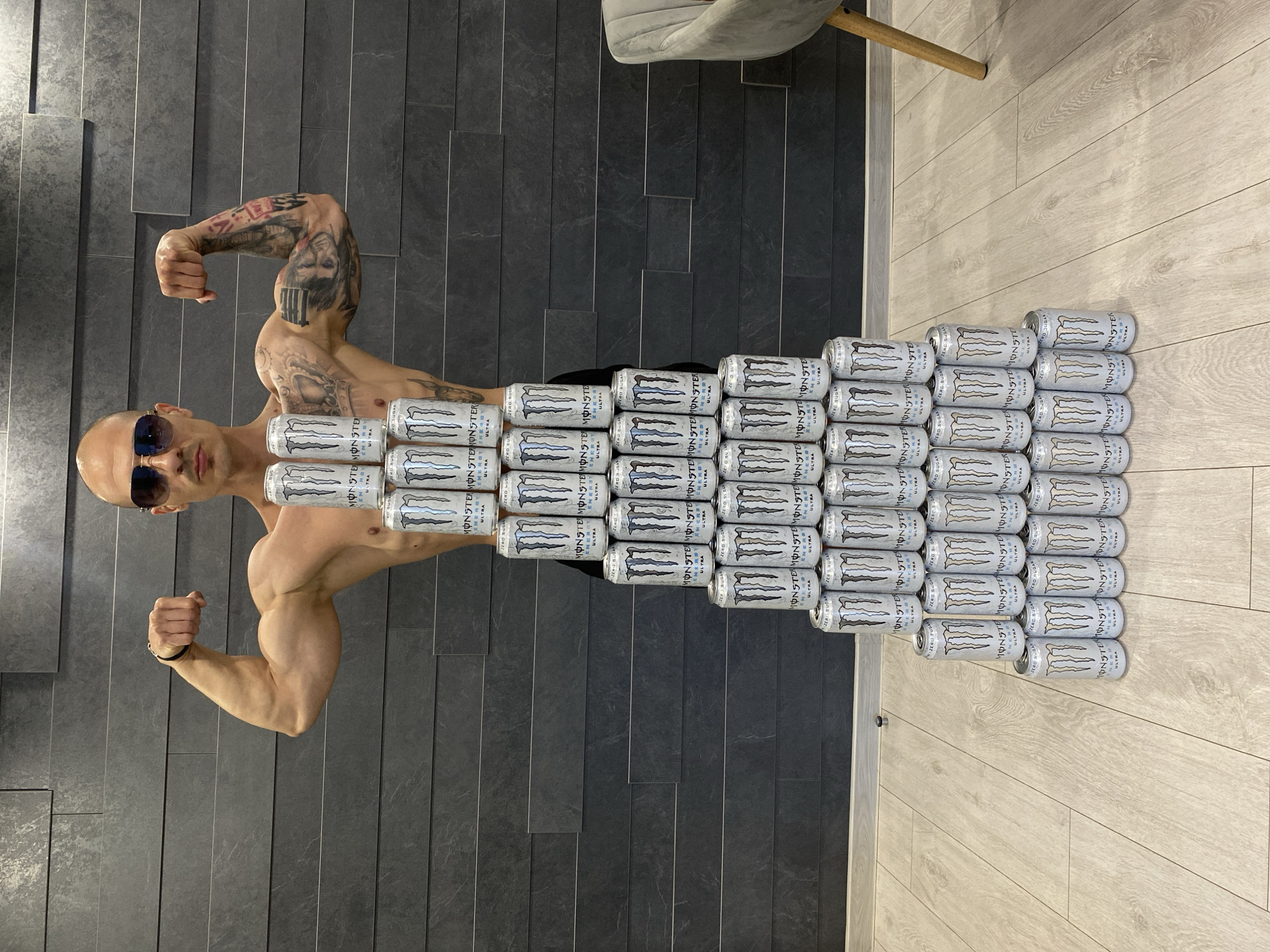 A muscular, bald man wearing sunglasses poses shirtless with flexed biceps against a dark gray brick wall. He is centered in the frame, with his body aligned vertically. Covering the man's torso and part of his legs is a stacked arrangement of about 45 cans of white Monster Ultra energy drinks. The floor transitions from dark gray brick to light beige wood. The leg of a chair is visible in the upper right corner. The location is not specified as Pleven, Bulgaria, in the image.