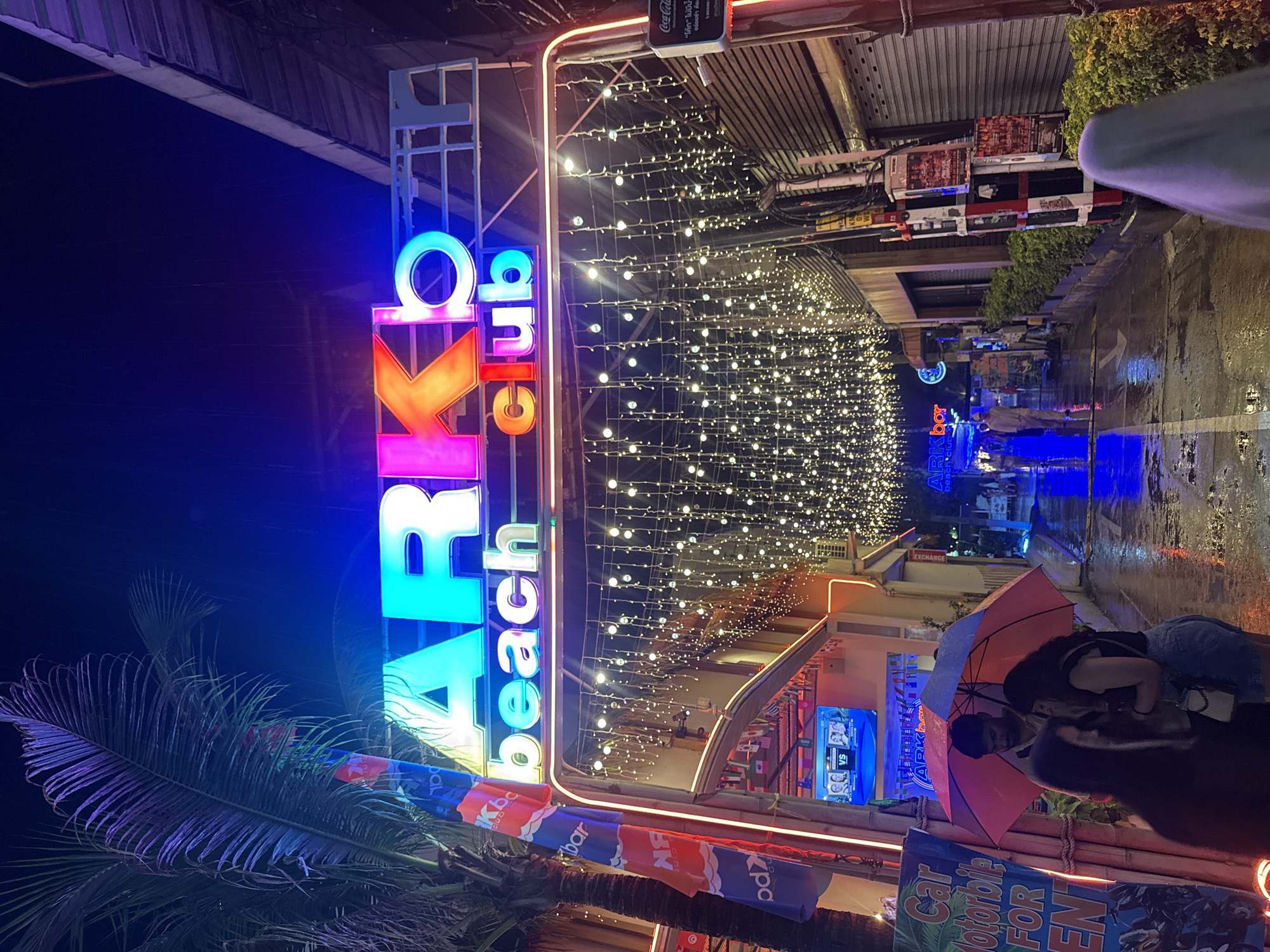 The image depicts a street scene at night in Baan Chaweng, Thailand, likely during or shortly after a rain shower, as indicated by the wet ground reflecting the neon lights. Dominating the upper portion of the frame is a large, brightly lit neon sign that reads "ARK beach club," with "ARK" in a mix of cyan and pink, and "beach club" in smaller, glowing letters below. A palm tree is visible to the left, its fronds illuminated by the colorful lights.
The street is lined with buildings, some with closed shutters, and adorned with strings of small, white fairy lights that create an archway over the walkway. A few individuals are present. In the foreground, three people are huddled under a large umbrella. To the left of them, a sign reads "Car Motorbike FOR RENT". Further down the street, other pedestrians are visible, walking away from the camera. The overall atmosphere is lively and typical of a tourist area at night.