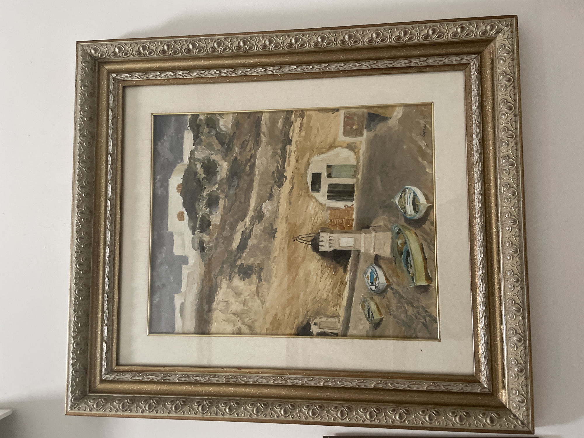 This is a framed oil painting depicting a coastal scene with several small boats pulled up on a sandy area. In the background, there are buildings carved into a hillside and a small white tower with a lantern at the top. The sky is overcast. The painting appears to be signed "Capra 95" in the lower right corner. The frame is ornate, with a distressed gold finish and carved detailing. The painting is hanging on a white wall.