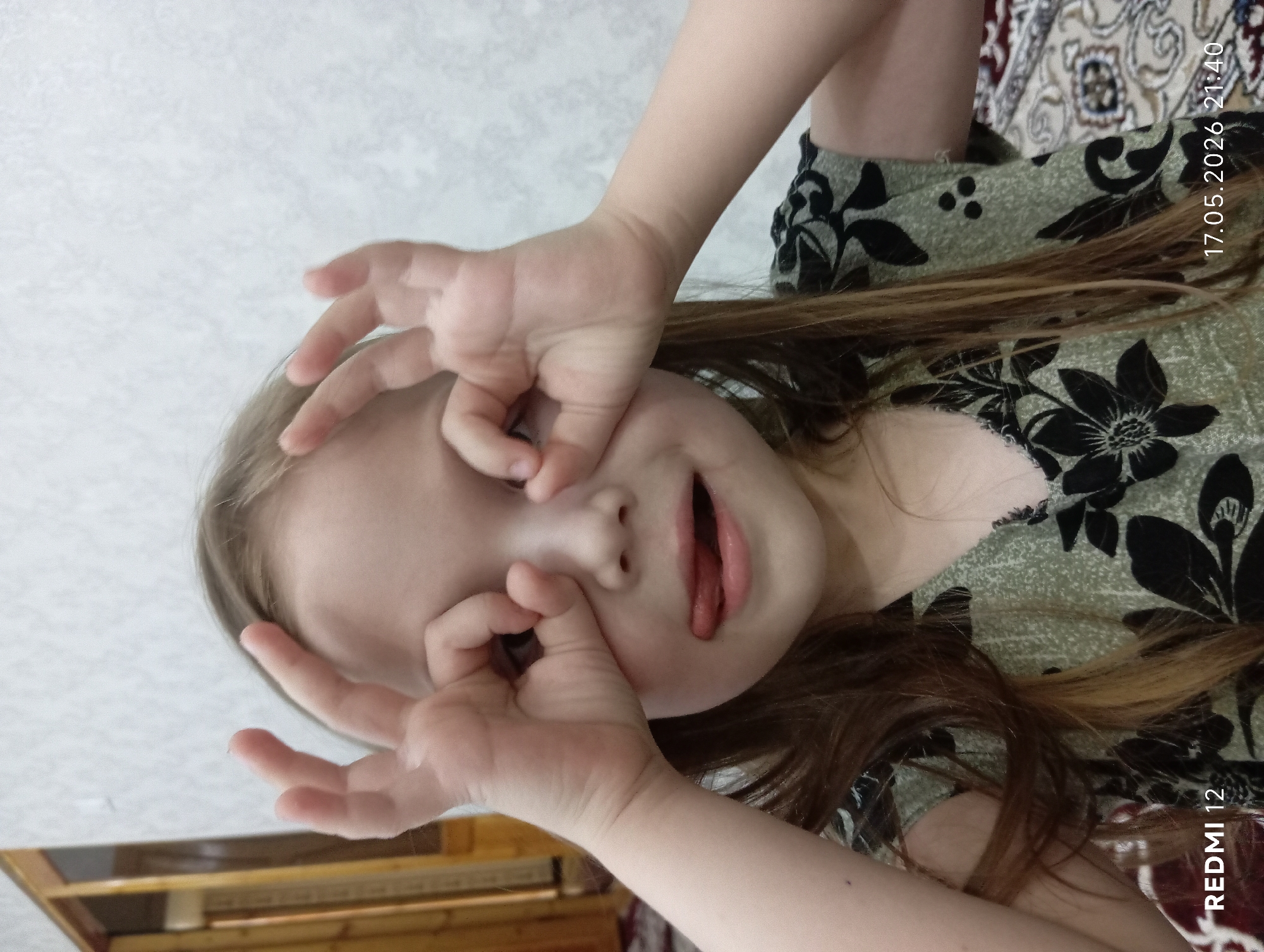 A young girl is posing for a picture. She is making circles around her eyes with her fingers and sticking her tongue out. She has light brown hair and is wearing a green dress with black floral patterns. The picture was taken on 17.05.2026 at 21:40, and the bottom right of the picture says "Redmi 12". The background behind her includes a patterned rug, and what appears to be a light-colored wall. It is not possible to identify the location as Bekabad, Uzbekistan from the image alone.