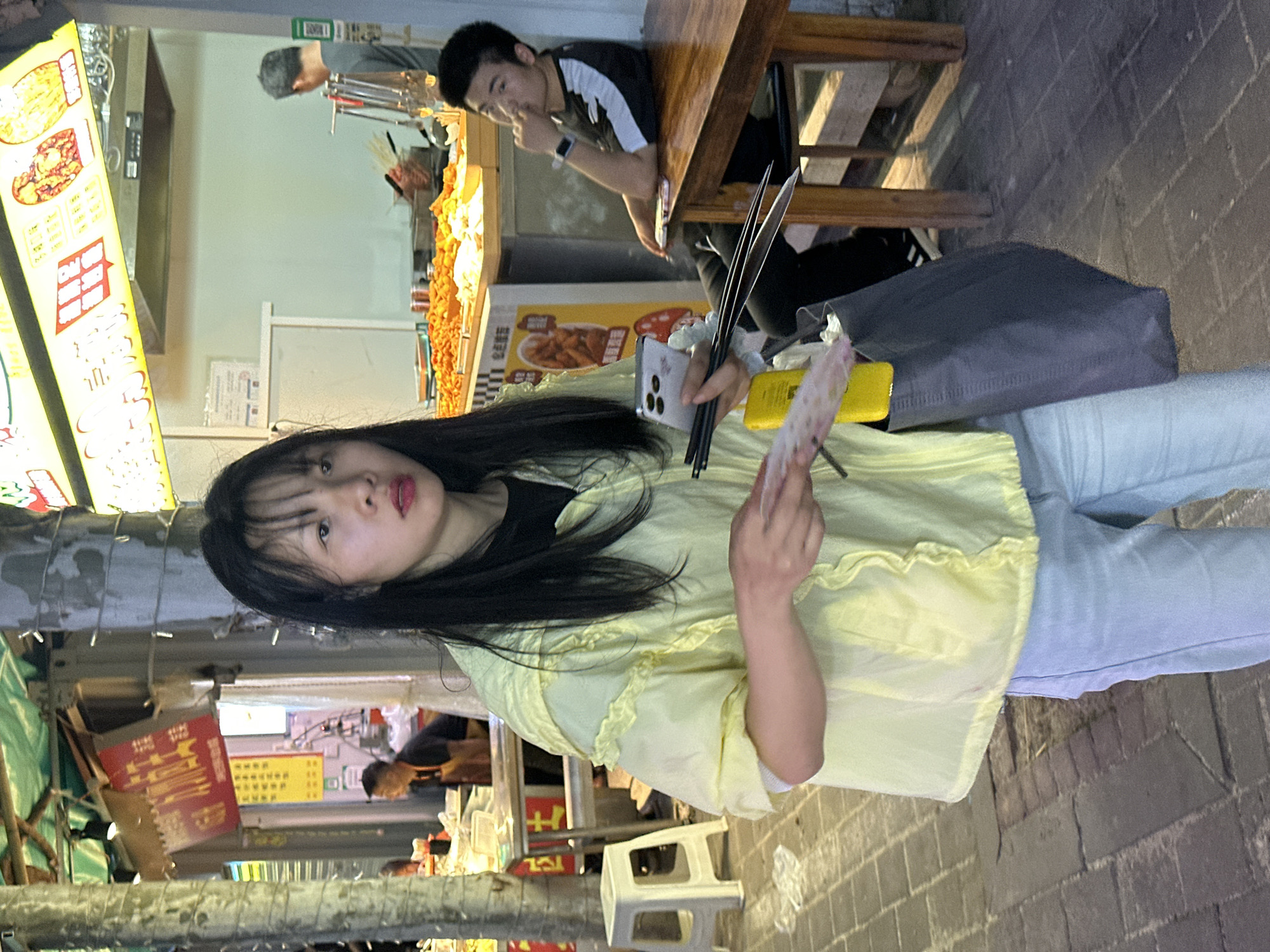 In Jiangjin, China, a woman with dark hair, wearing a yellow shirt and light blue jeans, holds chopsticks and money in her hands while standing on a brick-paved street near a food stall. The stall has illuminated signs displaying food items. A man is seated at a wooden table in the background, and there is a grey bag on the ground near the woman. The setting appears to be an outdoor food area, possibly at night given the lighting.