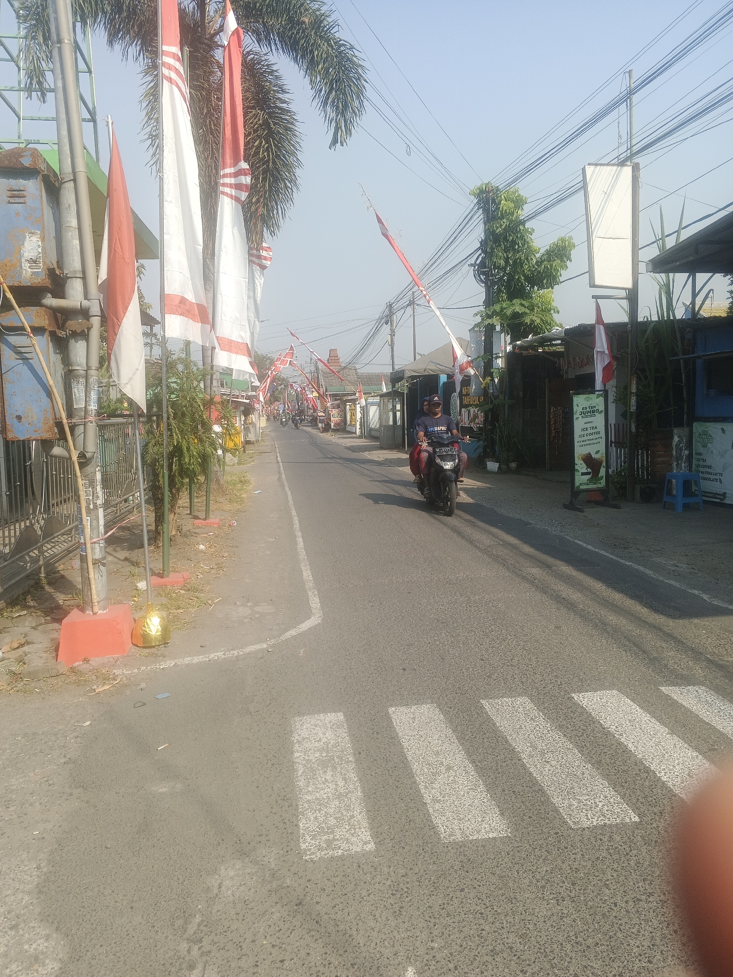 The image captures a street scene in Krian, Indonesia, with a focus on a paved road lined with buildings and pedestrian areas. The Indonesian flag, red and white, is prominently displayed on several poles, suggesting a national celebration or commemoration. A few trees and utility poles with a network of overhead wires are also visible.
In the foreground, a pedestrian crossing is marked by white stripes on the asphalt. On the right side of the road, a blue plastic stool sits near a building with a sign indicating drinks like "ES TEH JUMBO," "ICE TEA," and "ICE COFFEE." Another sign nearby lists various tea and coffee options.
Further down the road, a person is riding a motorcycle with another passenger. The motorcycle has a license plate that appears to read "L 3117 YYY." The buildings along the street exhibit various architectural styles, with some appearing to be shopfronts or small businesses. The sky is clear and bright, indicating a sunny day. The overall atmosphere suggests a typical day in a Indonesian town.