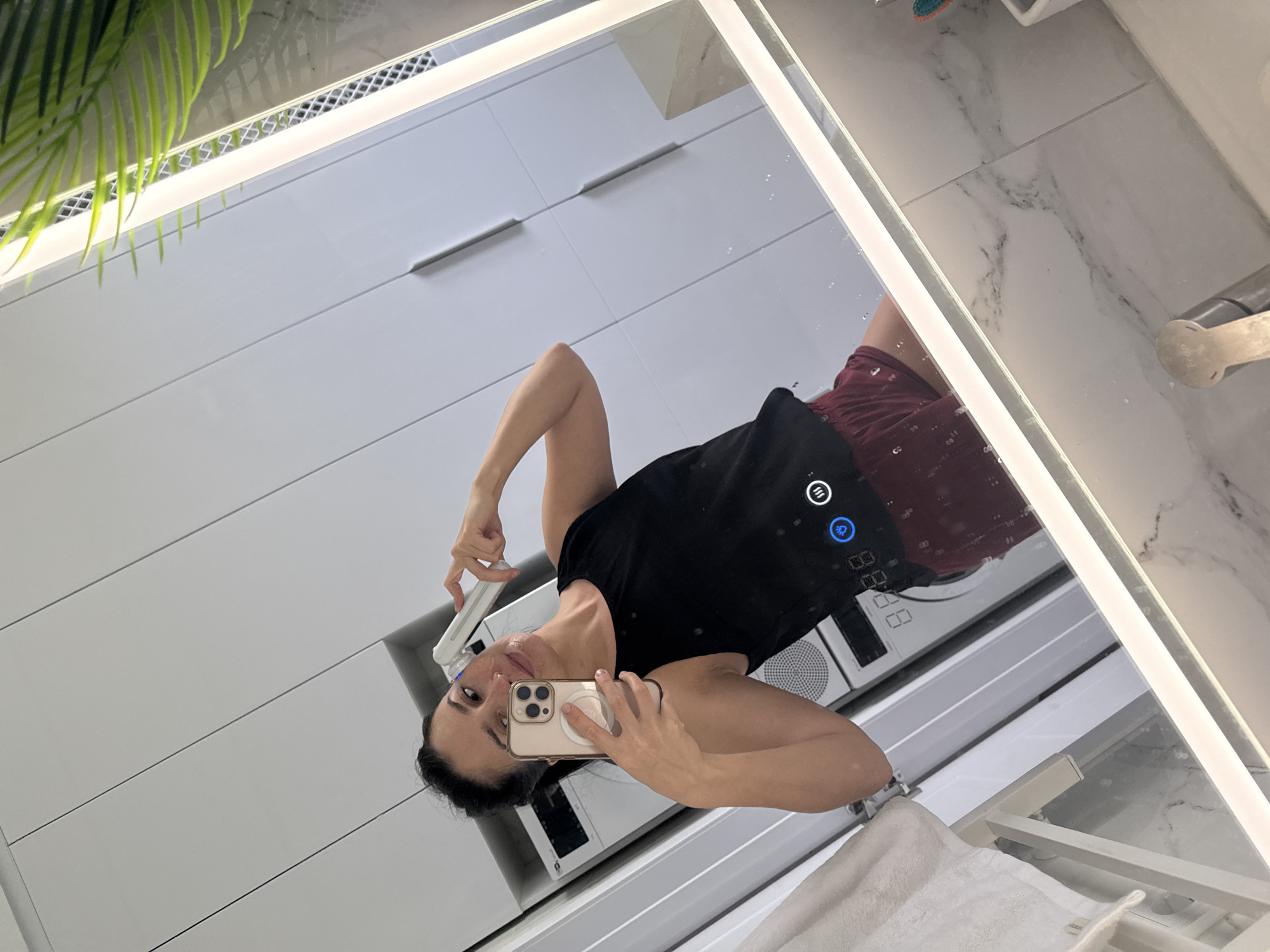 A woman is taking a mirror selfie in a modern bathroom. She is holding a smartphone in her left hand and a small white beauty device in her right hand, which she is using on her face. She is wearing a black tank top and maroon shorts. The bathroom has white cabinets, a large illuminated mirror, and marble-patterned tiles. A plant is visible in the upper right corner, and a towel hangs on a rack to the left. The scene appears to be in a residential setting.