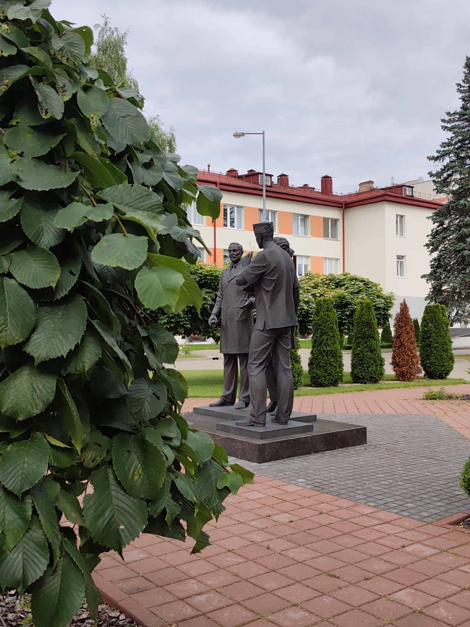 In a city park in Бараўлянскі сельскі Савет, Belarus, a bronze statue depicts three men in formal attire engaged in conversation. The statue is positioned on a square granite base. The foreground is dominated by a lush, green tree with large leaves, partially obscuring the left side of the monument. A paved walkway with red and grey brick patterns leads to the statue, forming a geometric design on the ground. In the background, a multi-story building with a red roof and cream-colored walls stands, interspersed with windows. A light pole is visible to the left of the building. A line of evergreen trees, some taller and some shorter, with varying shades of green and a single reddish-brown one, runs along the edge of the paved area. A mature fir tree stands to the right of the building, adding to the scene's arboreal elements. The sky is overcast and cloudy.