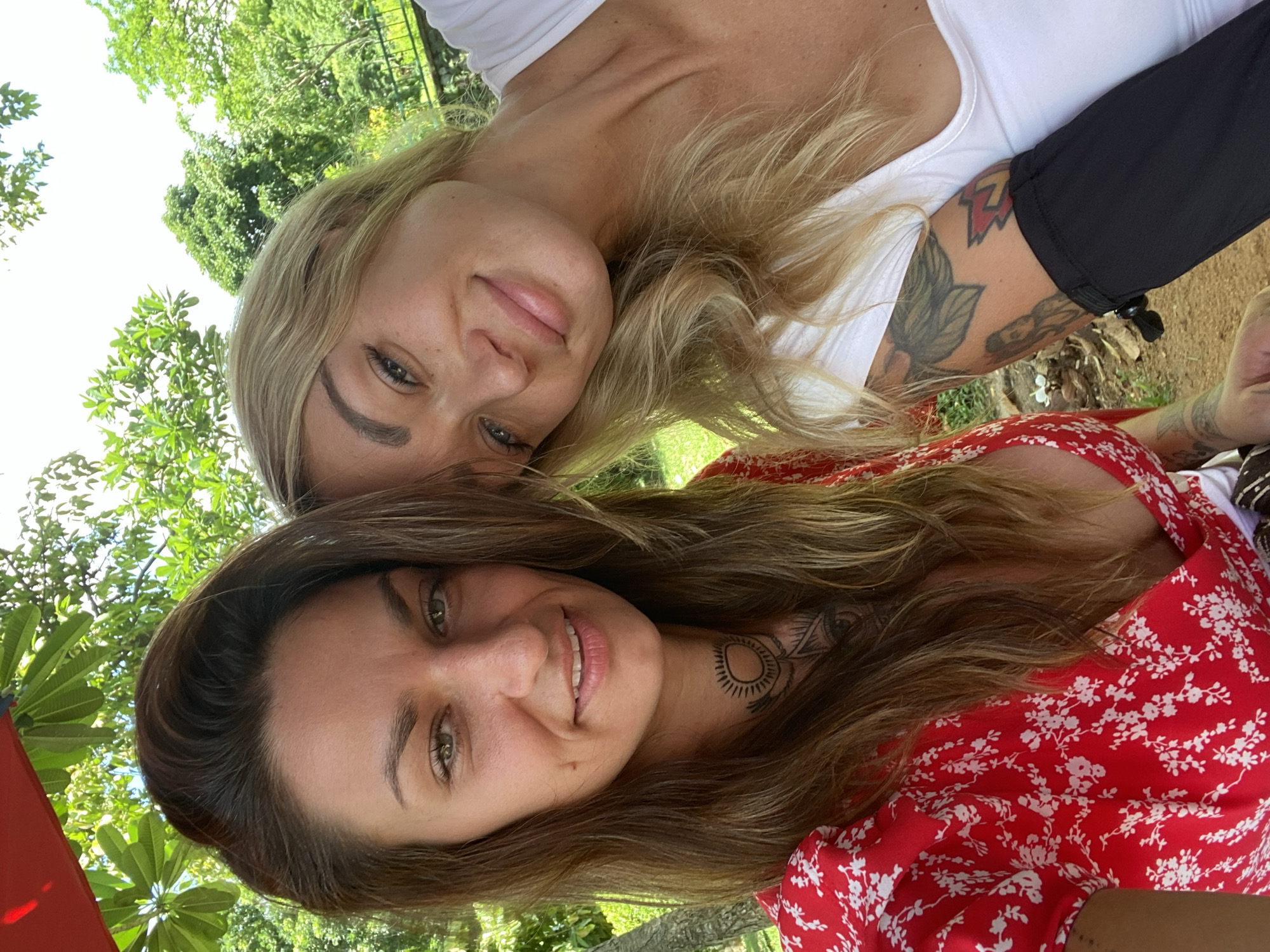 Two women are posing for a selfie outdoors. One woman has long blonde hair and is wearing a white top. Her left arm has tattoos. The other woman has long brown hair and is wearing a red floral print dress. She has a circular tattoo on her neck. They are positioned close together with trees and greenery visible in the background. The location is in Thủ Đức, Vietnam, based on the background scenery.