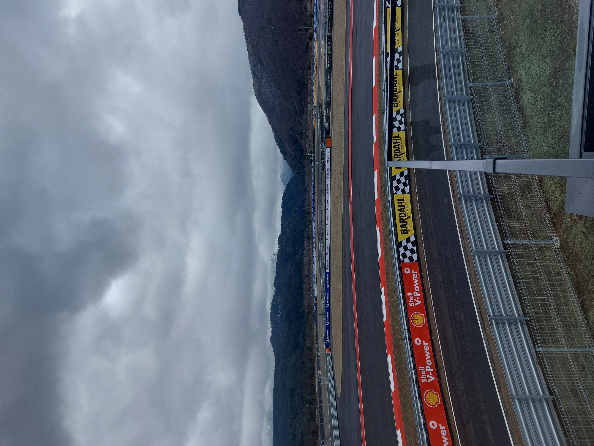 Here is a detailed description of the image:
The image shows a race track situated in an unknown location. There are multiple racing lanes and guardrails visible. Branding such as Shell V-Power, Bardahl, and Michelin are displayed along the trackside. In the background, a mountain range is partially obscured by a cloudy, overcast sky. A metal structure is also visible in the foreground.
