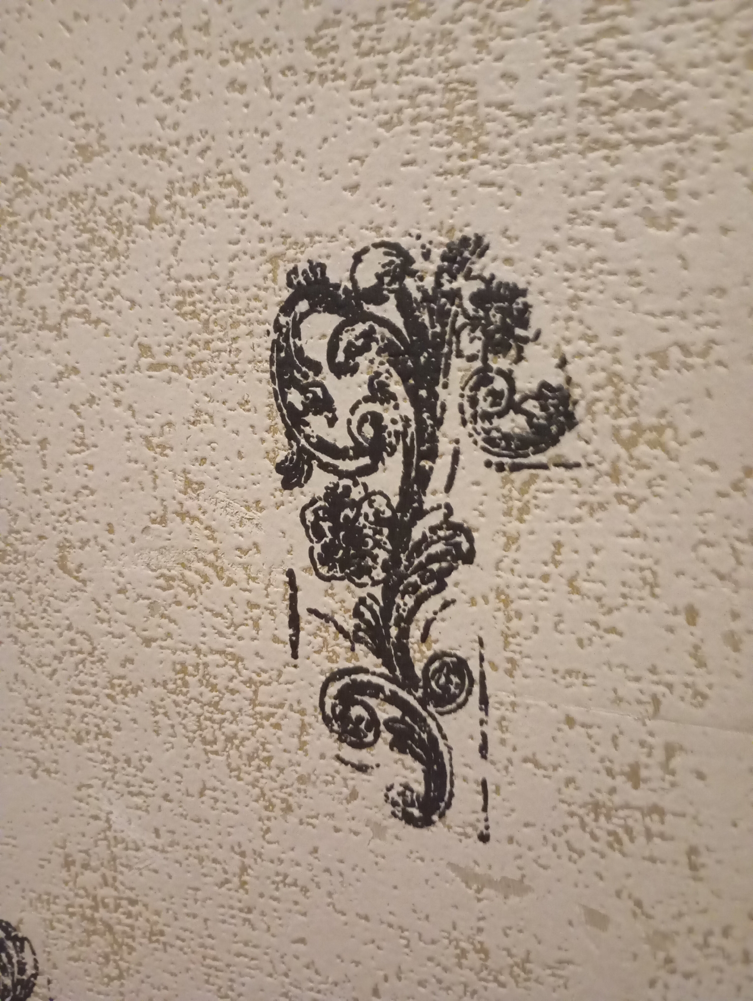 The image shows a close-up of a textured, beige-colored surface, possibly wallpaper or a painted wall. A black, ornamental stencil pattern is visible on the surface. The pattern is intricate, featuring floral motifs and swirling lines, resembling a stylized branch or vine with leaves and small circular elements. The texture of the surface creates a slightly uneven background for the stencil, giving it a raised or embossed appearance in some areas. The lighting casts subtle shadows, highlighting the texture of the wall and the depth of the stencil. The scene is indoors, with no indications of the surrounding environment. The location is Unknown, Unknown.