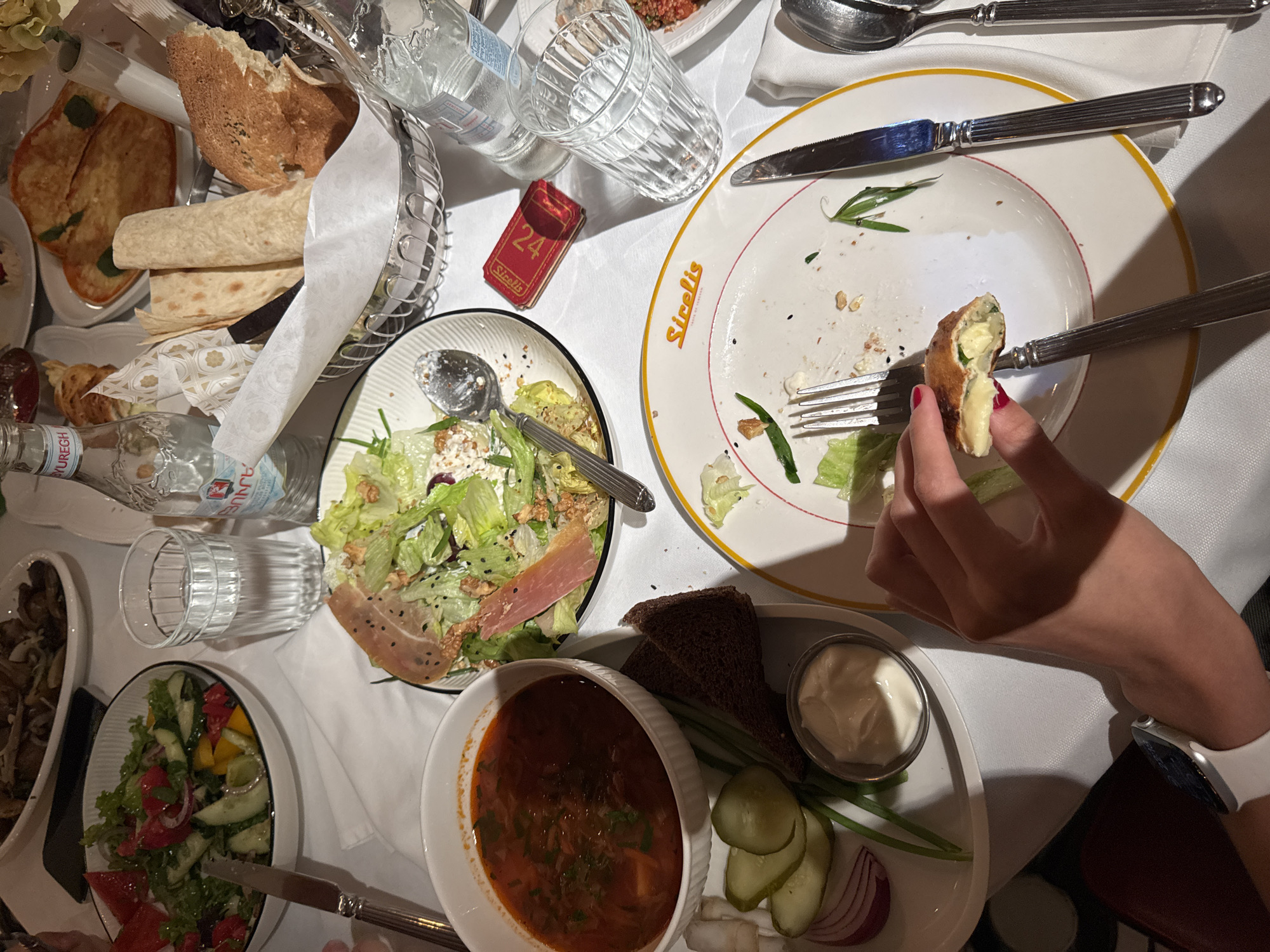 The image captures a table setting at a restaurant, seemingly in Yerevan, Armenia, based on the "Sirelis" branding on one of the plates. The table is covered with a white tablecloth and features a variety of dishes.
There's a bread basket with various types of bread, including lavash. Salads are visible, including one with lettuce, ham, and cheese, and another with cucumber, tomatoes, and red onions. A bowl of borscht is present, accompanied by sides of sour cream, dark bread, pickled cucumbers, red onion slices, and green onions.
Water is provided in glass bottles, one of which is labeled "Byuregh." There are also multiple empty drinking glasses. Silverware is scattered across the table. A small red box with the number "24" and the name "Sirelis" is positioned near the center of the table.
A person's hand is visible, holding a piece of food with a cheesy filling. The person wears an Apple Watch on their wrist.