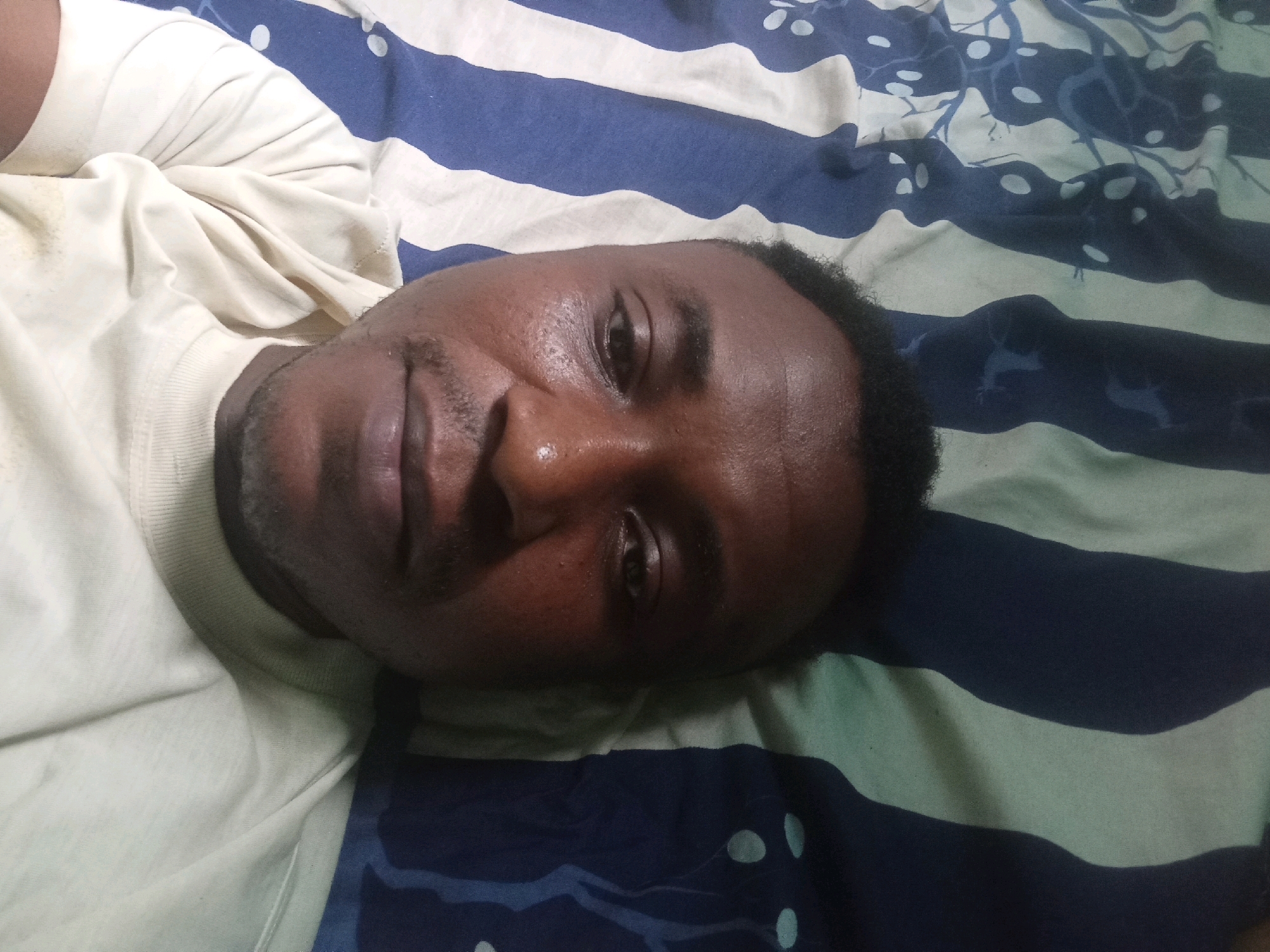 The image is a close-up, overhead, selfie-style shot of a man lying on his back on a bed with striped bedding. The man, who appears to be Black, is wearing a light-colored t-shirt. His head is slightly tilted to the right, and he is looking towards the camera with a slight smile. The bedding has alternating wide stripes of dark blue and light green, with a subtle pattern of white oval shapes and blue tree-like designs on the dark blue stripes. The lighting suggests an indoor environment, possibly a bedroom. The image does not contain any discernible landmarks or clues that would indicate its specific location within Agege, Nigeria.