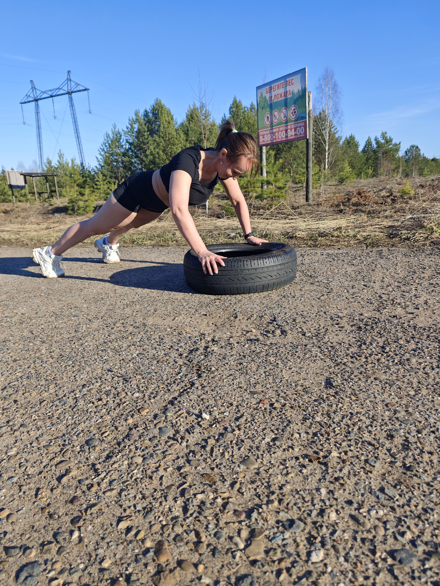 A woman is captured mid-exercise on a bright, sunny day, likely in Chaikovsky, Russia. She is performing a push-up or holding a plank position, with her hands placed firmly on a black car tire that rests on a gravel road. The woman has brown hair tied back and is dressed in black athletic attire, consisting of a short-sleeved top or sports bra and shorts, paired with white sneakers.
The setting is an unpaved road at the edge of a pine forest. In the background, tall green trees stretch under a clear, vibrant blue sky. To the left, a large metal power line tower is visible, along with a small wooden structure nestled among the trees. To the right, a white warning sign with a green top border stands tall. The sign displays Russian text: "БЕРЕГИТЕ ЛЕС ОТ ПОЖАРА" (PROTECT THE FOREST FROM FIRE). Below this, there are four circular symbols with red diagonal lines, indicating prohibitions against fire, picnics, smoking, and driving off-road. Further down, the sign lists "ПРЯМАЯ ЛИНИЯ ЛЕСНОЙ ОХРАНЫ" (FOREST PROTECTION HOTLINE) with the phone number "8-800-100-94-00" and states "БЕСПЛАТНО КРУГЛОСУТОЧНО" (FREE 24/7). The ground is composed of small stones and dirt, and strong shadows cast by the subjects and objects suggest bright daylight. The scene depicts outdoor fitness in a natural, open environment.