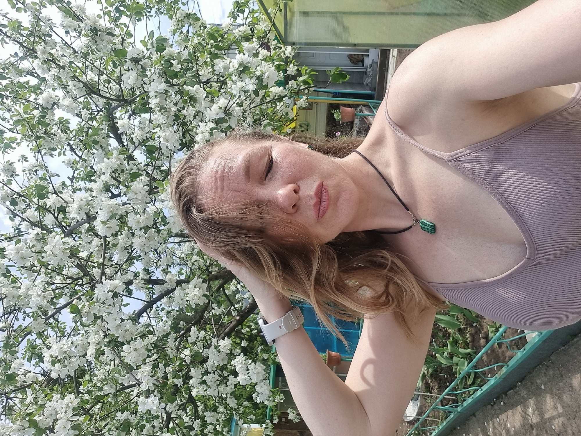 A woman is posing for a photo outdoors. She is fair-skinned with blonde hair. She is wearing a mauve-colored ribbed tank top and a necklace with a green pendant. She has a watch on her left wrist. Her eyes are closed, and she has a relaxed expression. In the background is a flowering tree with white blossoms. There is also a fence, a building with windows and potted plants visible. The photo was taken in the city of Chaikovsky, Russia.