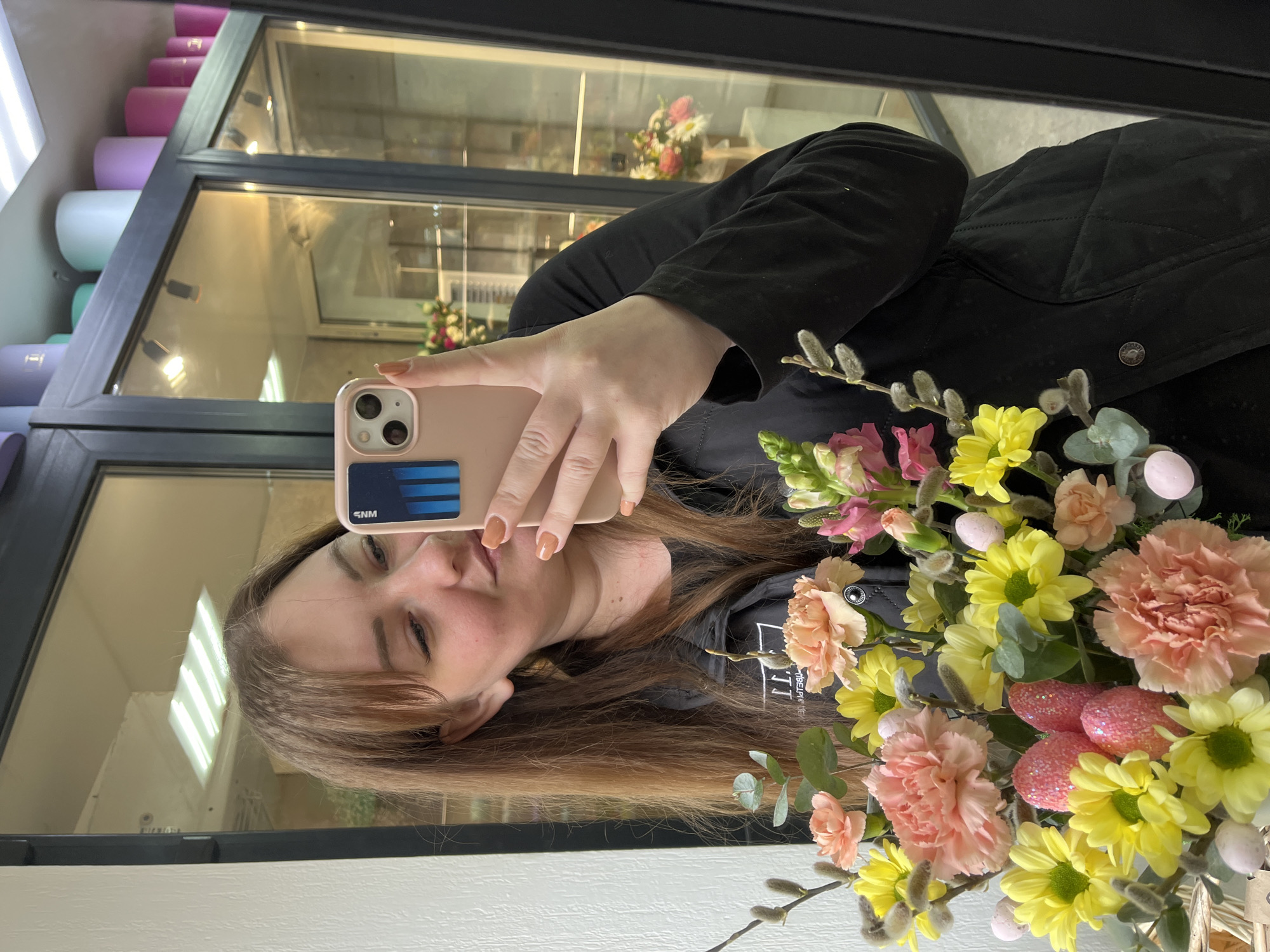 A person is taking a selfie with a bouquet of flowers in what appears to be a flower shop. The individual, who is of the female gender, has medium-length brown hair and is holding a smartphone with a peach-colored case. The bouquet features an assortment of flowers including pink carnations, yellow daisies, and pussy willow branches, interspersed with small pink decorative eggs. The person is wearing a black quilted jacket. The backdrop reveals shelves stocked with what appear to be storage containers of varying colors. Outside the window, reflections suggest a building across the street.
The location is inferred to be in the city of Chaikovsky, Russia, based on the phone case sticker.