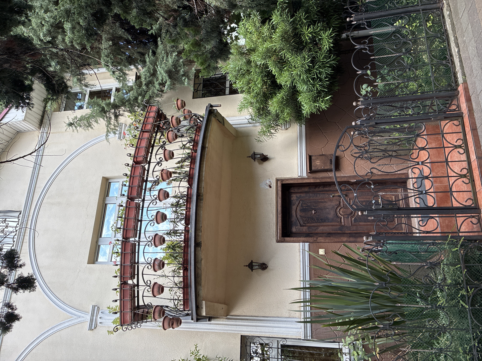 The image shows the facade of a building in Sochi, Russia, with a balcony adorned with potted plants and an ornate metal railing. A dark wooden door is visible on the ground floor, flanked by an outdoor lantern and a mailbox. A decorative wrought iron fence encloses the property, with lush green vegetation and trees surrounding the entrance. The architecture suggests a residential or hotel property, typical of the coastal city's style.