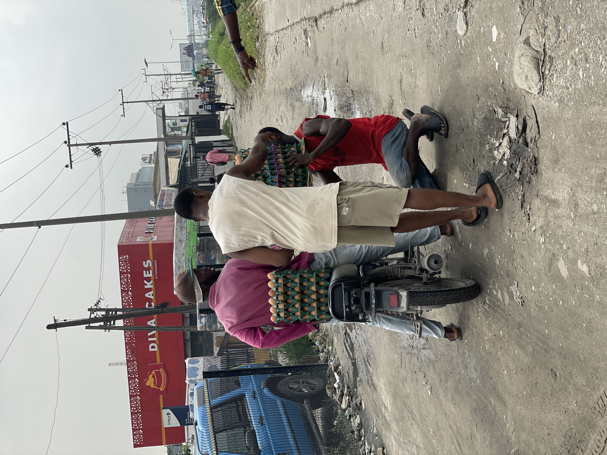 In Igboefon, Nigeria, on a cloudy day, three men are gathered around a motorcycle loaded with egg cartons. The man on the left, wearing a pink shirt and a brown hat, is seated on the motorcycle. Another man in a white tank top and tan shorts stands to the right of the motorcycle. A third man in a red tank top and blue jeans is kneeling and holding two stacks of egg cartons. In the background, a blue SUV is parked behind a fence, and the red building of "DIVA CAKES & Confections" is visible. The scene unfolds on a dirt road with scattered debris, and several utility poles with overhead wires line the street. Other pedestrians can be seen in the distance walking along the road.