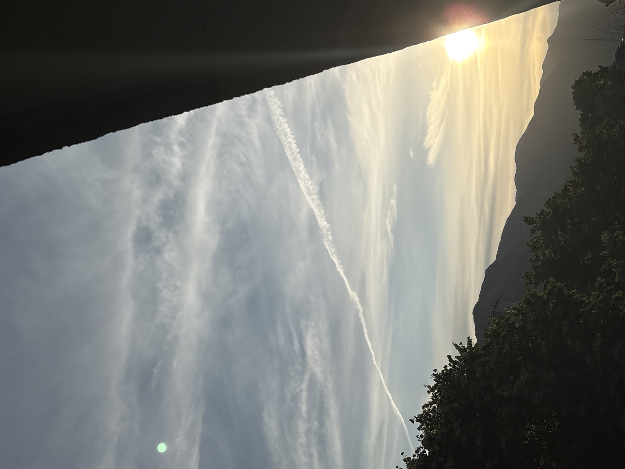 The image shows a sky filled with wispy clouds during what appears to be sunset or sunrise. The sun is a bright, warm glow near the top of the frame, casting a golden light that illuminates the clouds and the silhouette of a mountain range on the right. A distinct contrail from an airplane cuts diagonally across the sky. In the foreground on the left, a dark, angled structure partially obscures the view, suggesting an overhang or architectural element. On the far right, the dark silhouette of dense trees is visible against the bright sky, with a hint of some distant man-made structures. A small, bright green circular anomaly is present in the lower left quadrant of the image, possibly a lens flare or reflection. The scene evokes a sense of calm and natural beauty, with the warm light and dramatic cloud formations creating a visually striking natural landscape. The overall atmosphere is peaceful and serene, capturing a moment of transition between day and night.