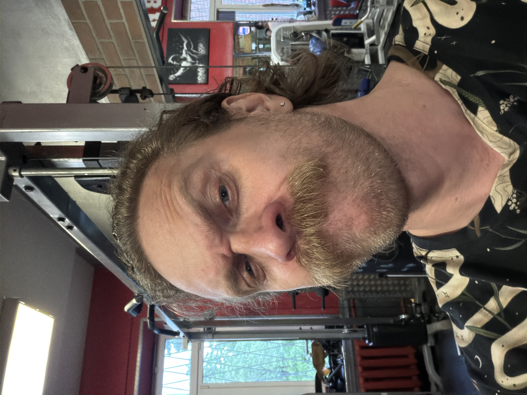 A selfie taken indoors, likely in a home gym. A Caucasian male with blondish-brown hair, a mustache, and a beard is the subject. He's wearing a dark shirt with a light-colored pattern. Gym equipment surrounds him. A poster hangs on a brick wall. A window is visible in the background, showing a view with trees outside. The location is not determinable as Saint Petersburg, Russia.
