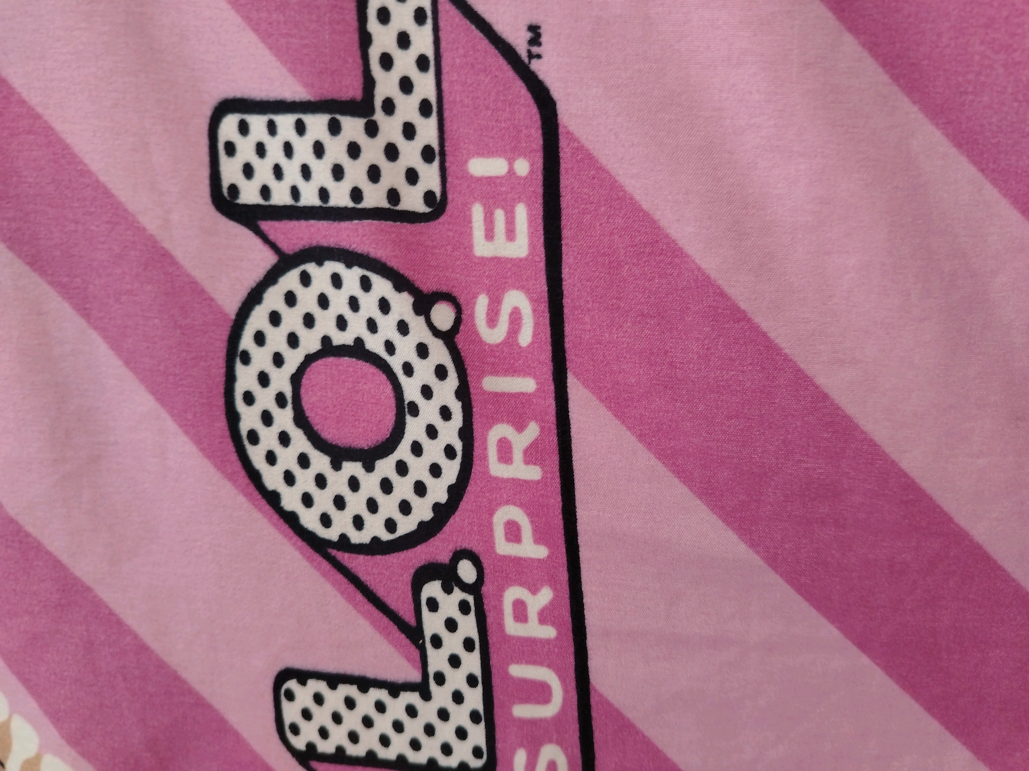 The image displays a close-up, angled shot of a pink fabric with diagonal stripes. Prominently featured is the text "LOL SURPRISE!" in large, bold lettering. The letters "L", "O", "L" are outlined in black and filled with black polka dots on a white background. The word "SURPRISE!" is written vertically in white capital letters with black outlines. A small trademark symbol (™) is visible above and to the right of the "L" in "LOL". The fabric appears to be soft and textured, possibly a blanket or clothing item. The background consists of alternating pink and lighter pink diagonal stripes. The image does not provide any information about the city of Чавусы, Belarus, nor does it depict any people, actions, or interactions.