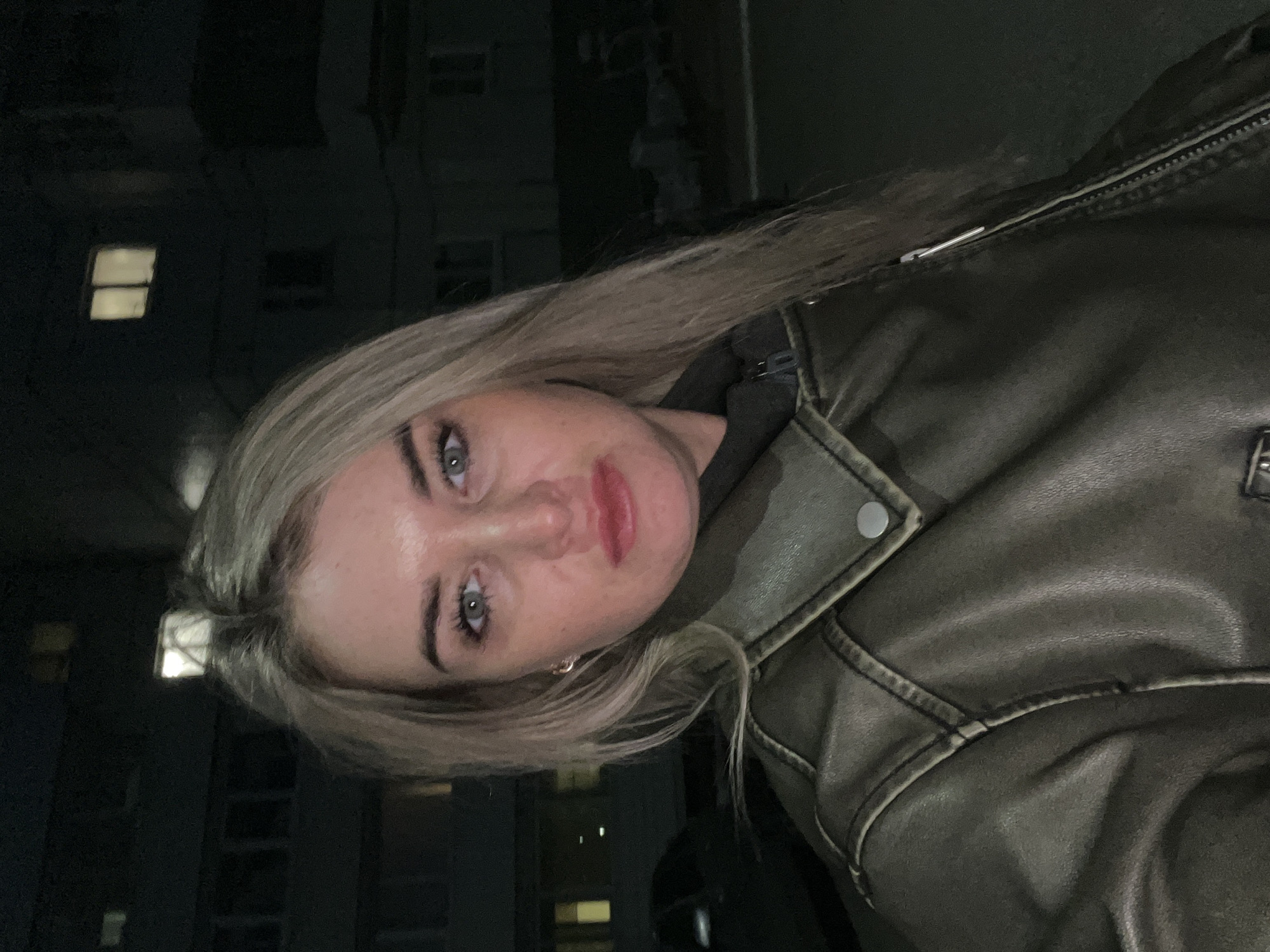 A woman is pictured at night in what appears to be the city of Chaikovsky, Russia. She has blonde hair, fair skin, and blue eyes and wears a brown leather jacket over a dark hooded sweatshirt. The background features buildings with lit windows. The image is taken from a close perspective, emphasizing the woman's facial features and clothing.