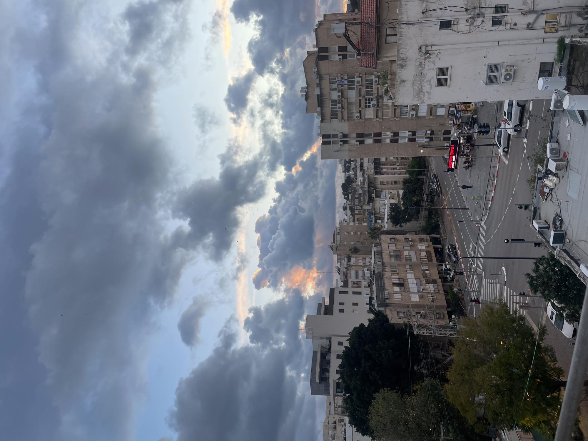The image presents an elevated urban landscape in Petah Tikva, Israel, captured during twilight or early evening. The sky is predominantly covered by extensive, dark grey cumulus clouds, interspersed with areas of lighter blue. A distinct, vertically oriented shaft of orange-pink light penetrates a cloud formation in the mid-right, indicative of a setting sun. Multiple small, dark specks, consistent with birds, are visible against the cloudy sky.
The cityscape below features numerous multi-story residential and commercial buildings. A modern, light-colored building with a flat roof is situated on the left, partially obscured by a large, dark green tree. Towards the right, a visibly aged, light-toned building displays exposed external wiring and multiple air conditioning units; its rooftop contains white water tanks and HVAC systems. Further back, a beige multi-story building features what appears to be an Israeli flag draped from a balcony.
At street level, an active urban intersection is visible, delineated by white crosswalks and lane markings. Traffic lights are present, showing red signals facing the viewer. Several vehicles are on the roadway, including a white SUV/station wagon near the intersection's center and another white car closer to the foreground. Pedestrians are present on the sidewalks. On the right side of the street, a commercial establishment with a prominent red sign displaying Hebrew text (reading "SUPER FREYTS") offers outdoor seating where several individuals are gathered. Streetlights are illuminated, and overhead power lines are visible throughout the scene.