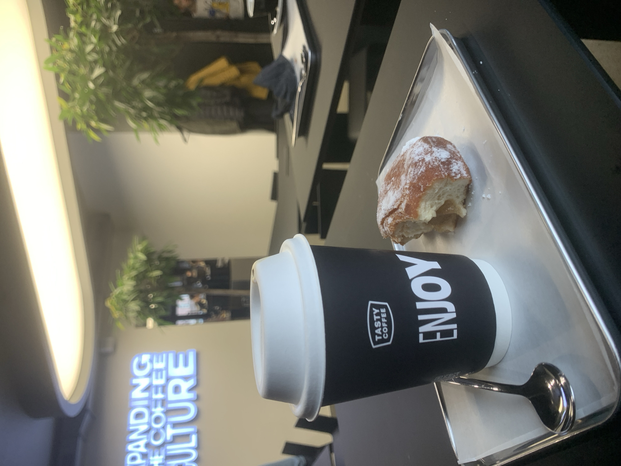 A close-up indoor shot on a tray with a coffee cup and pastry in a café or restaurant in Izhevsk, Russia. The coffee cup is black with a white lid and the word "ENJOY" printed vertically on its side, along with the logo of "TASTY COFFEE." Beside the cup is a silver spoon and a pastry covered in powdered sugar, with a bite taken out. The tray is metallic with a white paper liner. In the background, out of focus, are plants, a sign reading "XPANDING THE COFFEE CULTURE," and elements of the café's interior, including tables and chairs.