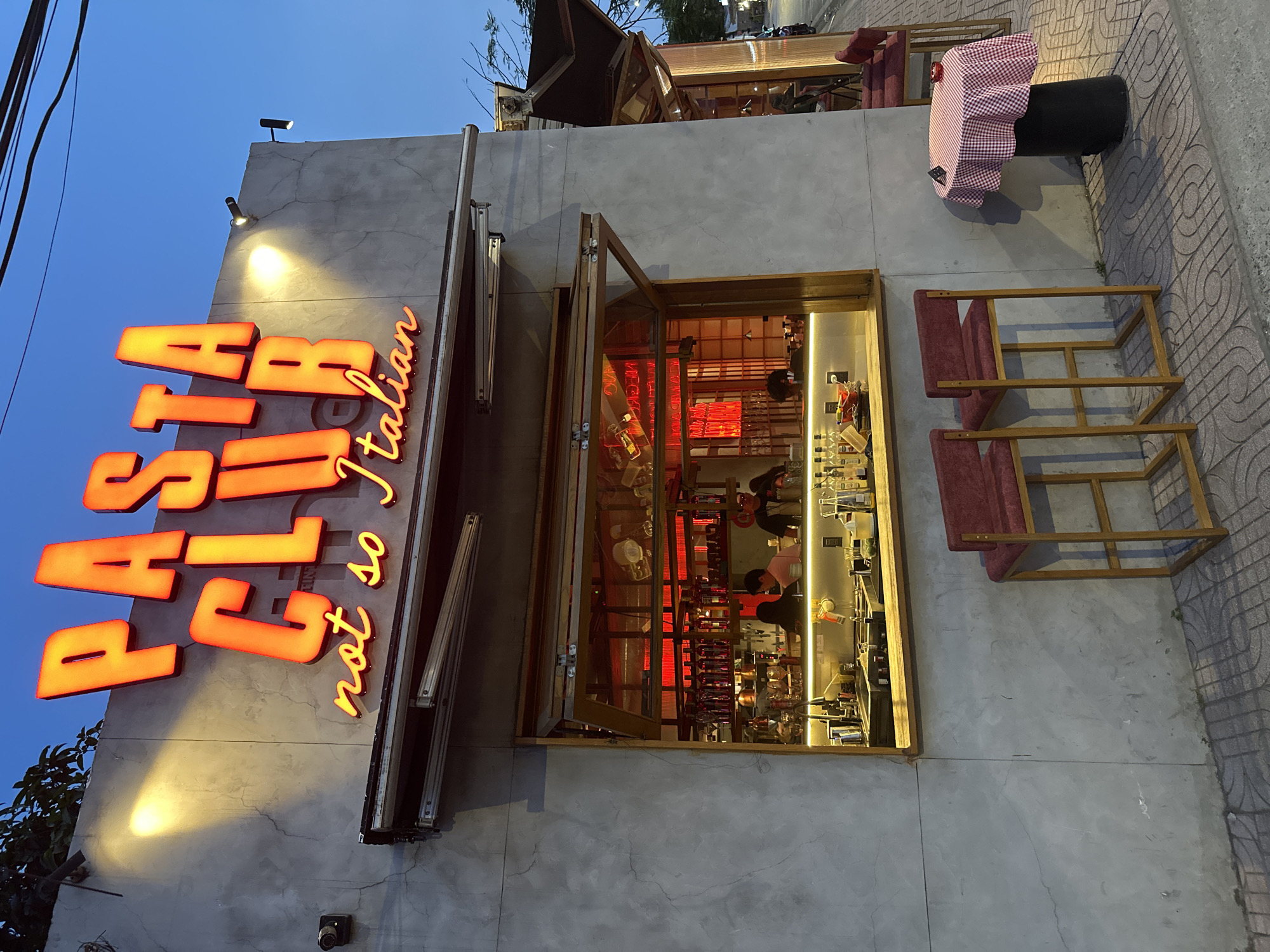 Here is a description of the image:
A ground-level view of a restaurant or bar exterior. A large neon sign reading "PASTA CIA" and "not so Italian" vertically adorns the left side of the facade. A wide window is centered, providing a view into the interior, which is illuminated and contains bottles, bar equipment, and at least two people at the counter. Red lighting elements are also visible within the interior. To the right of the window, two wooden chair frames with red seats are positioned on the sidewalk. Further to the right, a small table with a black cylindrical base and a red and white checkered tablecloth stands on the sidewalk. The facade is concrete with visible cracks and discolorations. The setting is likely evening or dusk.
Based on information available online, "Pasta CIA" is a restaurant located in the city of Thủ Đức, Vietnam.